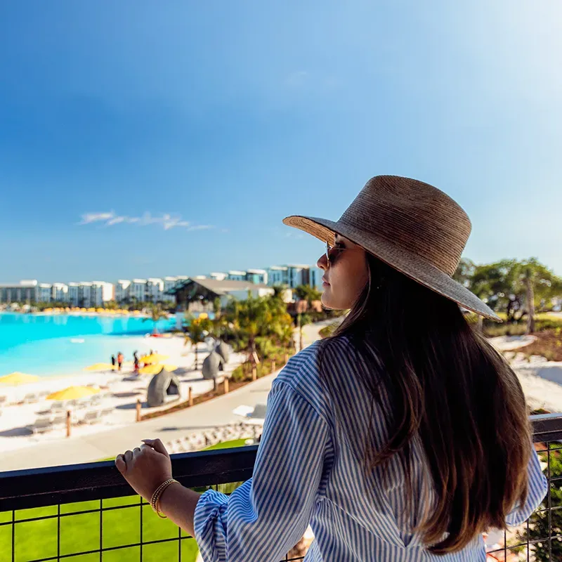 Woman in sunglasses and wide hat overlooking a resort pool on a sunny day.