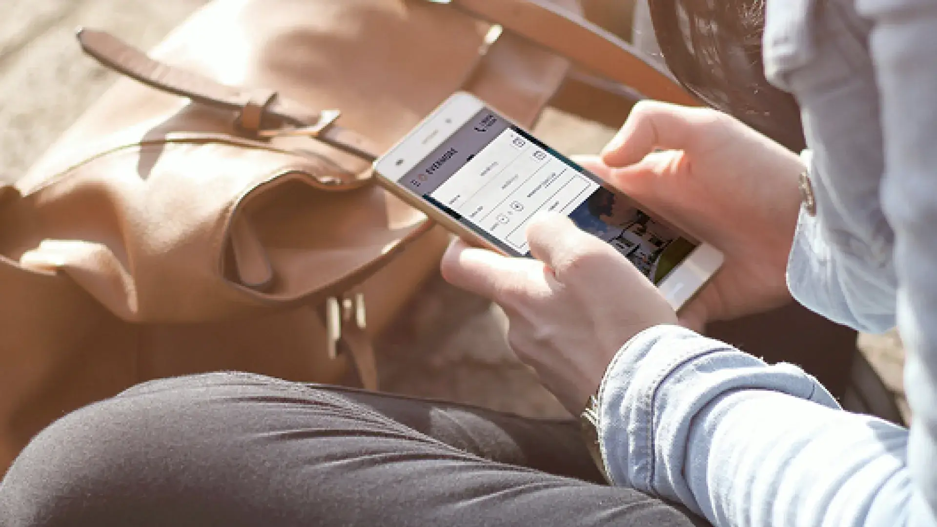 Person holding a smartphone showing a travel app interface near a tan leather bag outdoors.