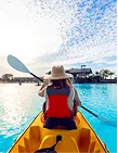 Person in hat and red life jacket paddling a yellow kayak on calm water under a blue sky.