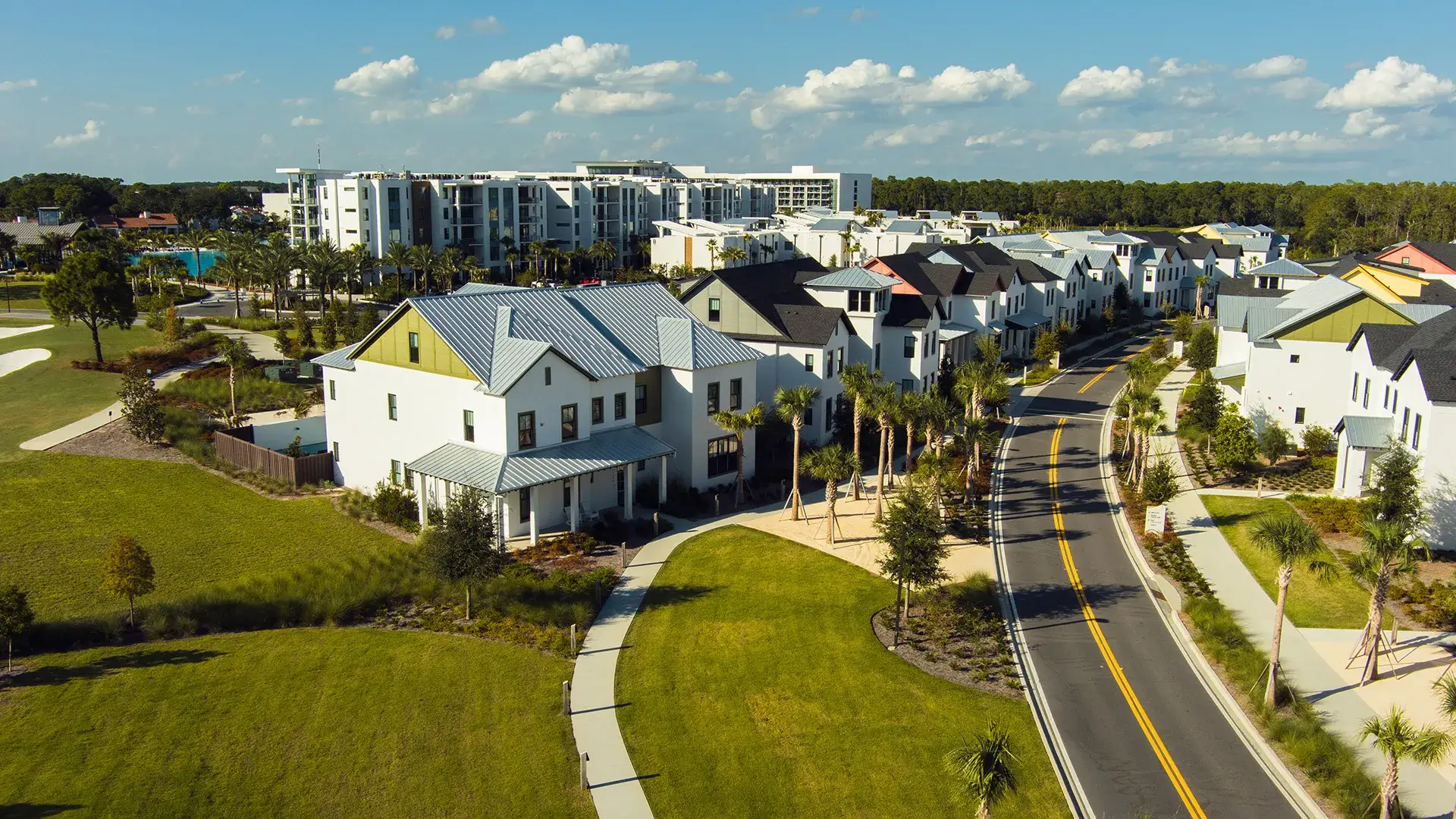 Aerial view of a suburban neighborhood with white houses, green lawns, and a winding road under a blue sky.