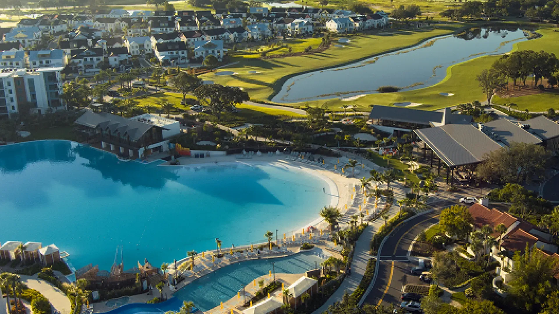 Aerial view of a resort with a large pool, beach area, golf course, and nearby residential buildings.