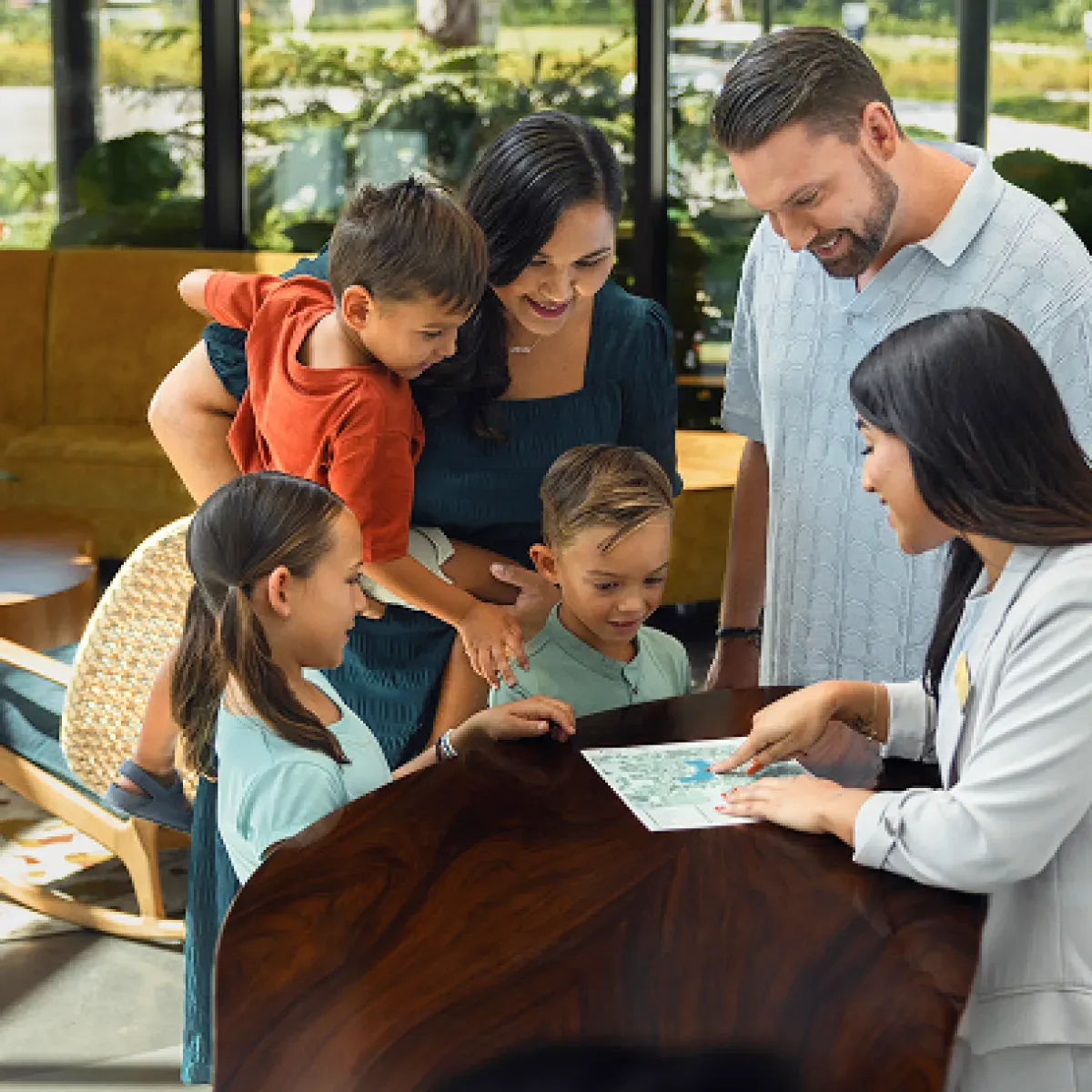 Family of five with two kids at a table while a woman in a white coat points to a map or brochure.
