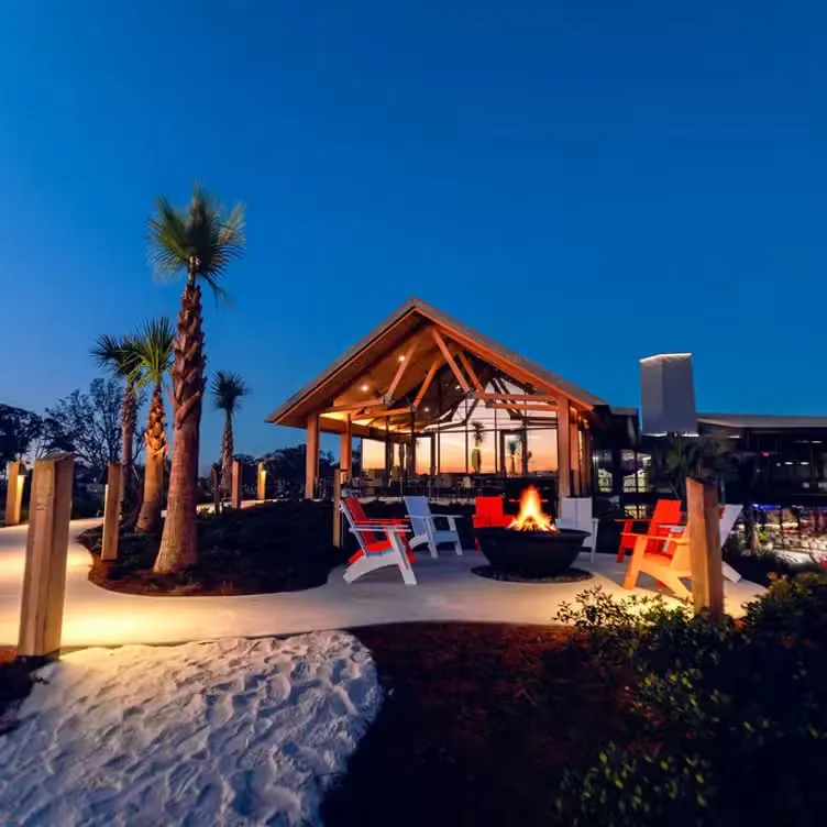 Outdoor fire pit with red and white chairs under a wooden pavilion at dusk with palm trees nearby.