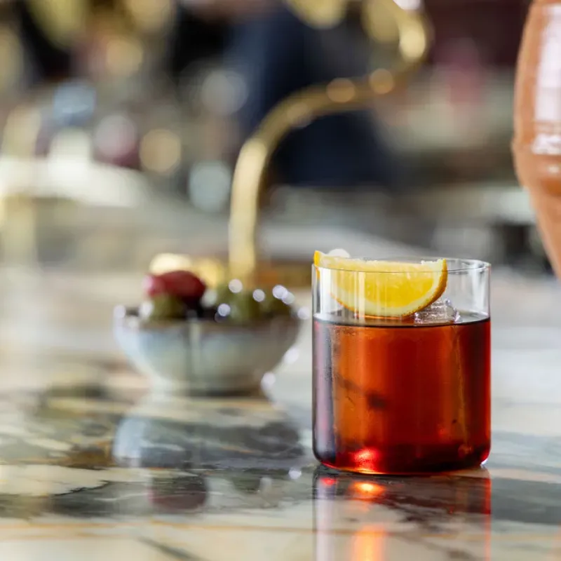 Glass of red cocktail with ice and an orange wedge on a marble counter with a bowl of olives in the background.