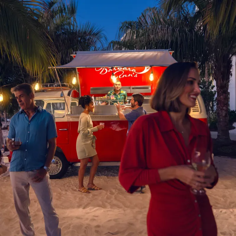 People enjoying drinks at a lively beachside bar in a red vintage van at dusk.