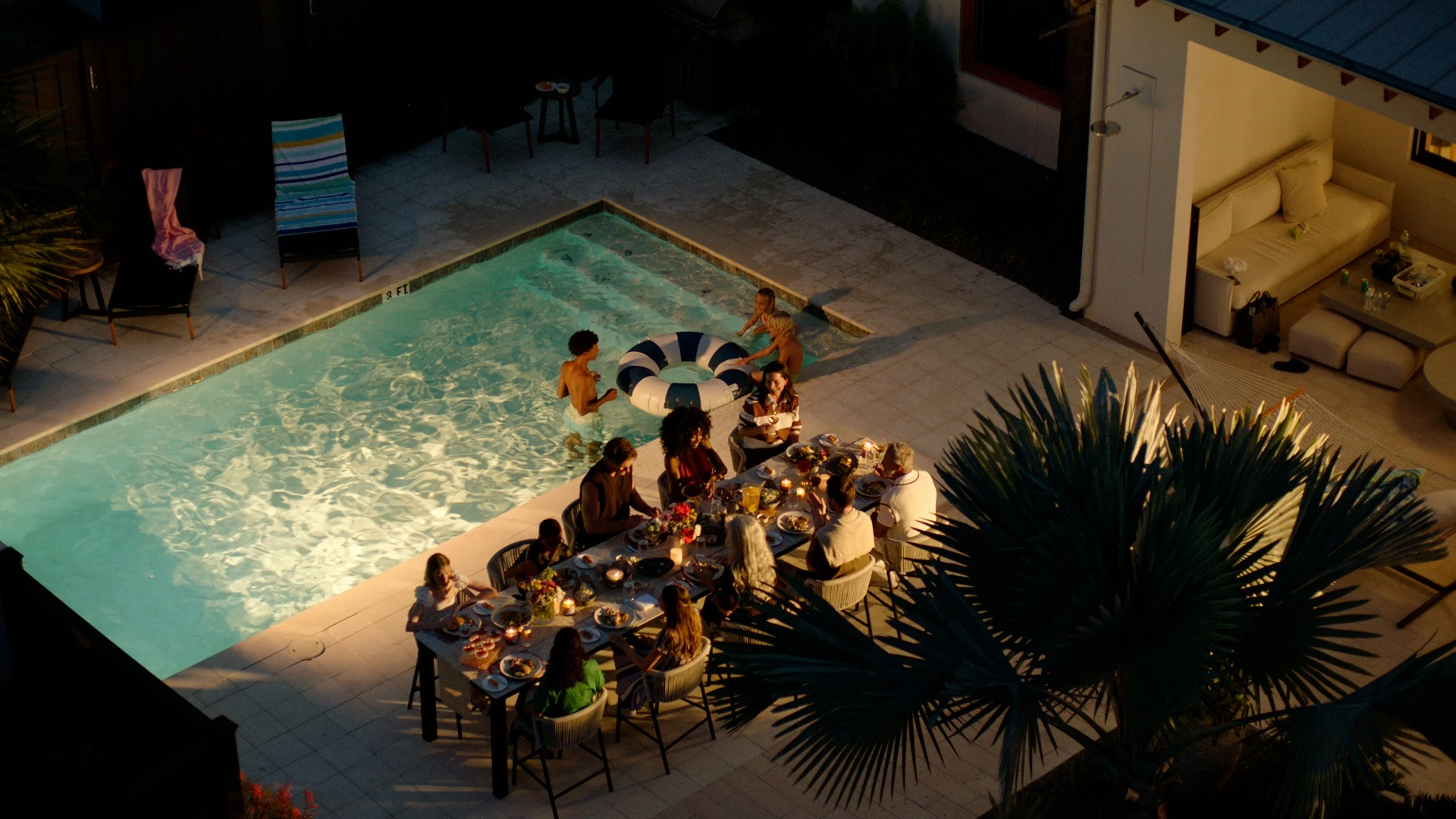 Group of people dining beside a lit swimming pool with two children playing with a float in the water.