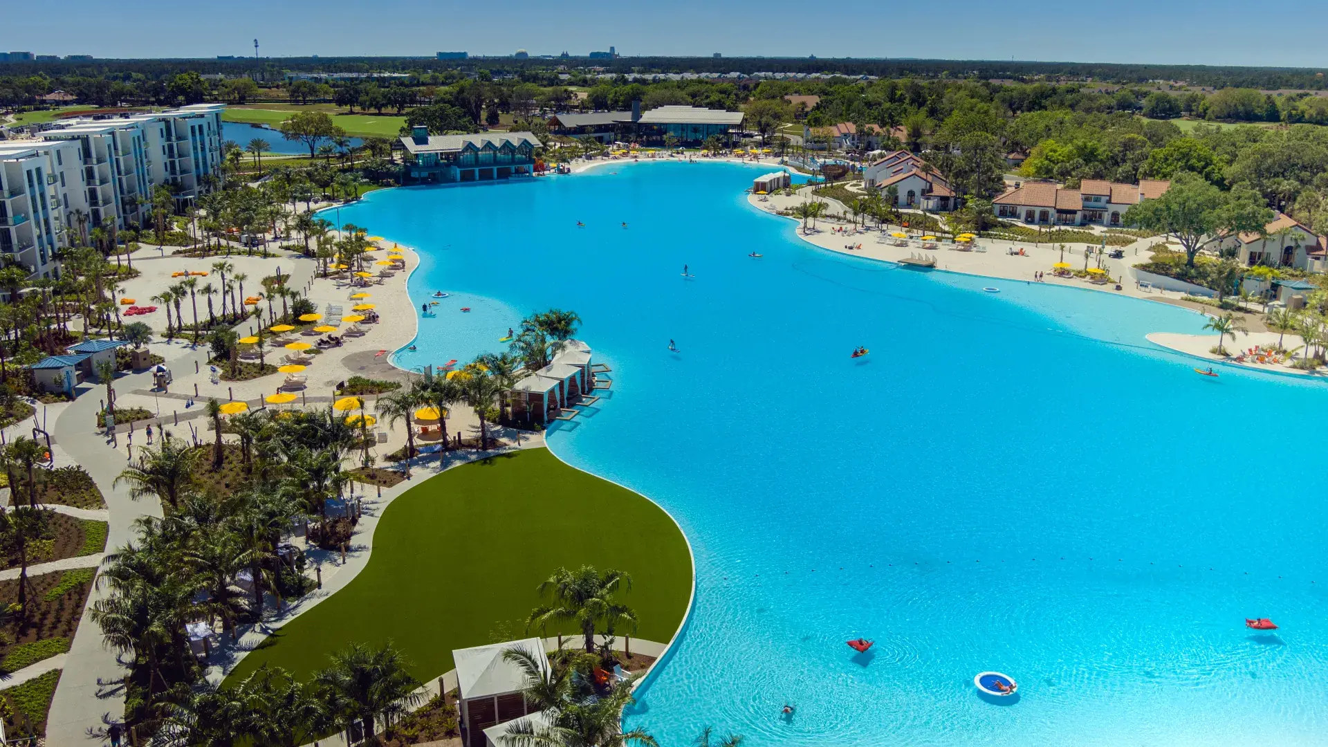 Large blue lagoon pool with people kayaking and standing, surrounded by palm trees and buildings.
