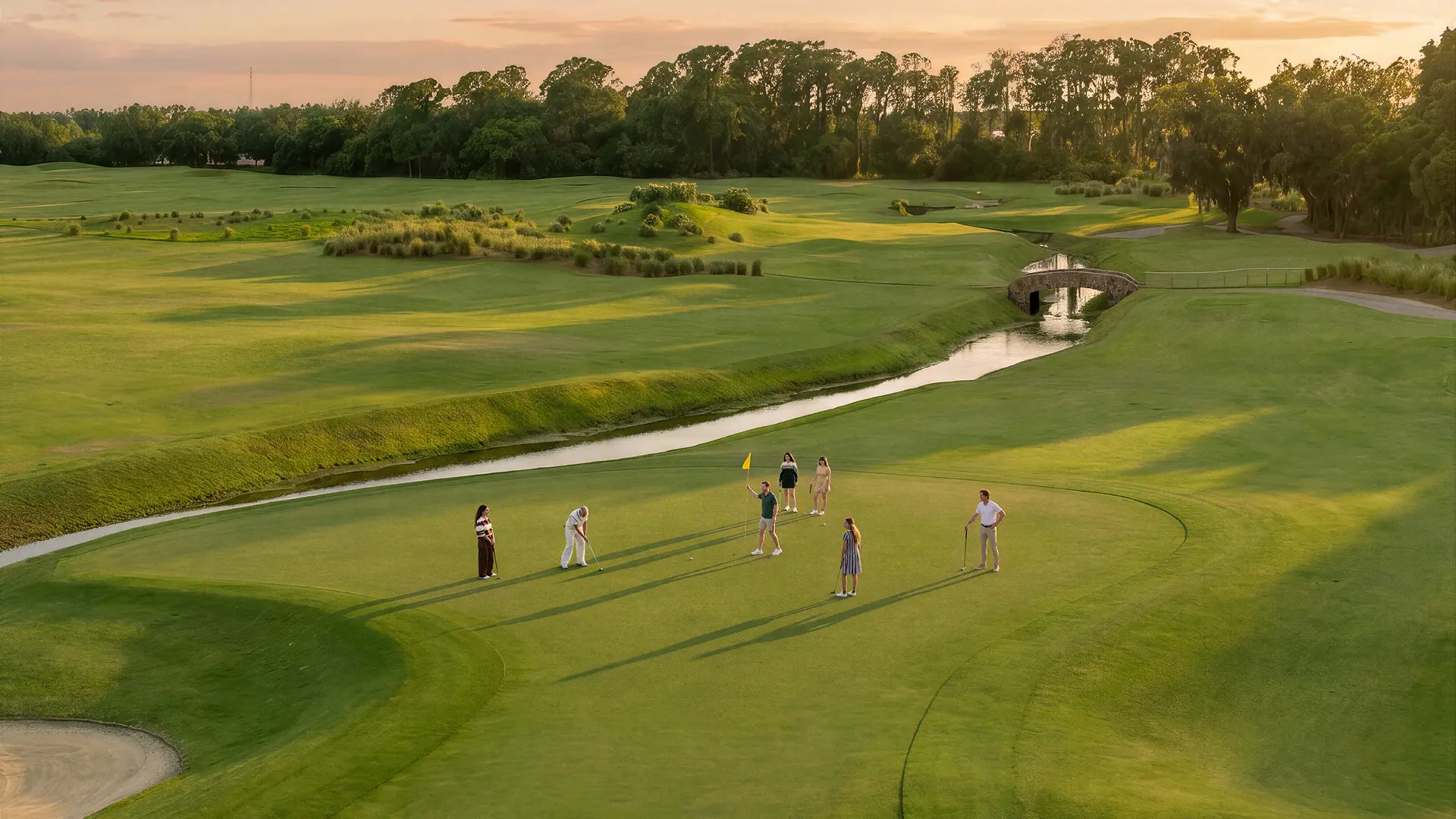 Six people on a golf course green with flagstick near a water hazard and trees at sunset.