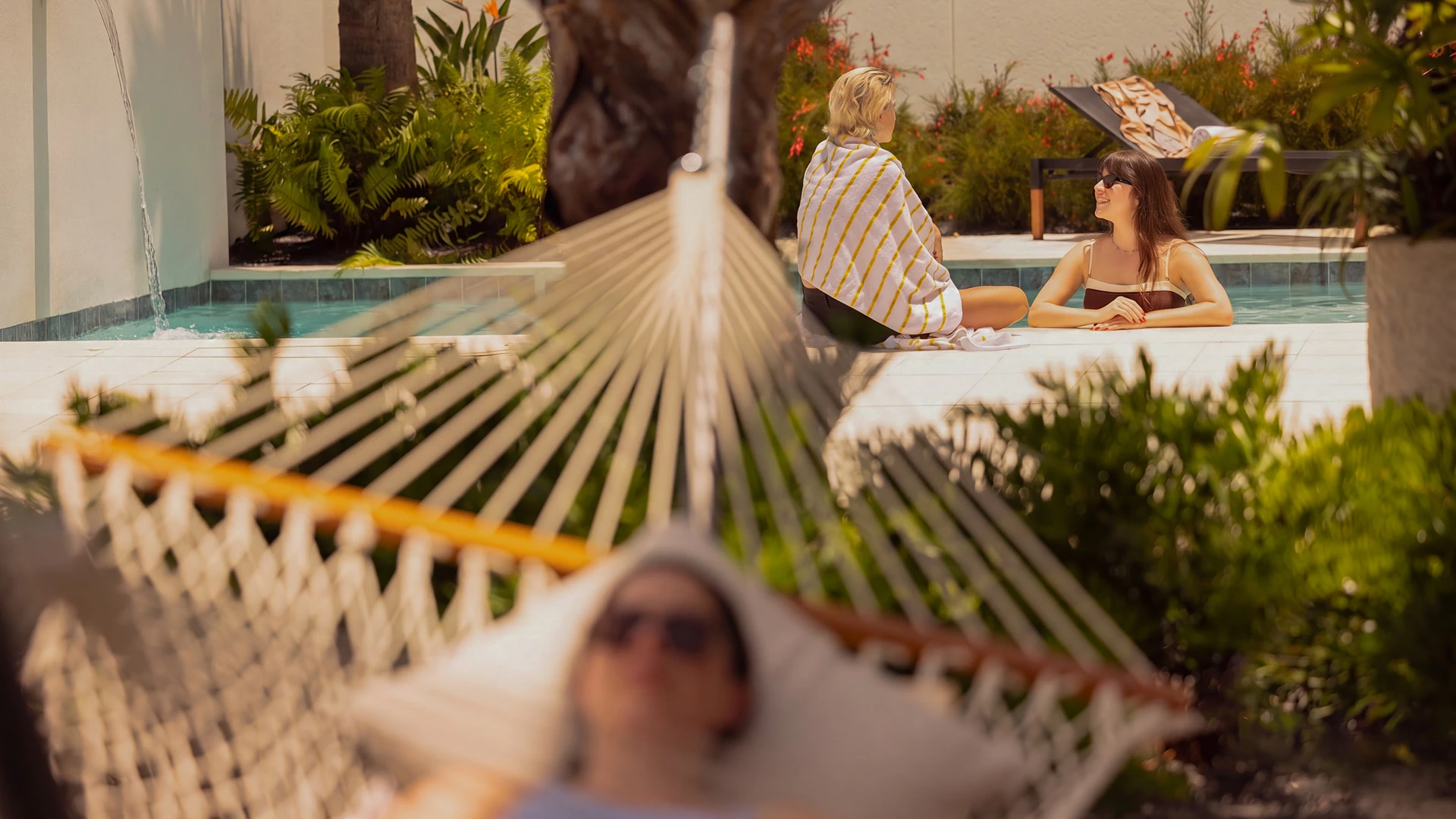 Two women talking at a sunny poolside while a person relaxes in a hammock in the foreground.