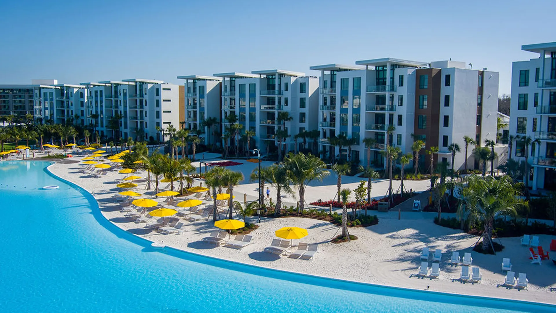 Large curving pool with yellow umbrellas and lounge chairs in front of modern apartment buildings.
