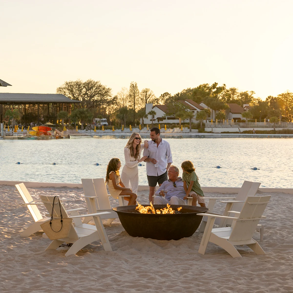 Family of five gathered around a fire pit on a sandy beach by a waterfront at sunset.