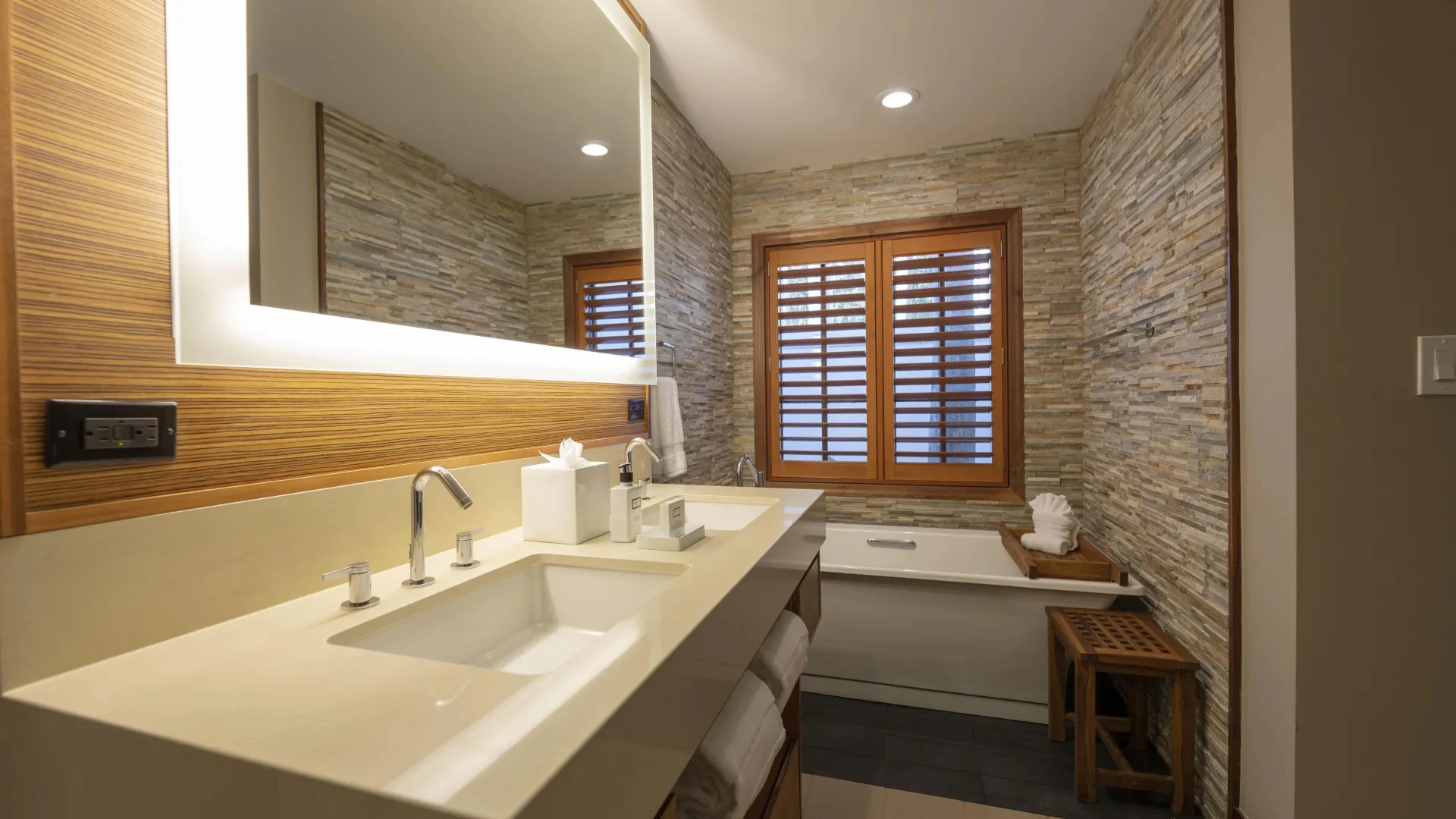 Modern bathroom with double sinks, illuminated mirror, stone wall, wooden window shutters, and bathtub.