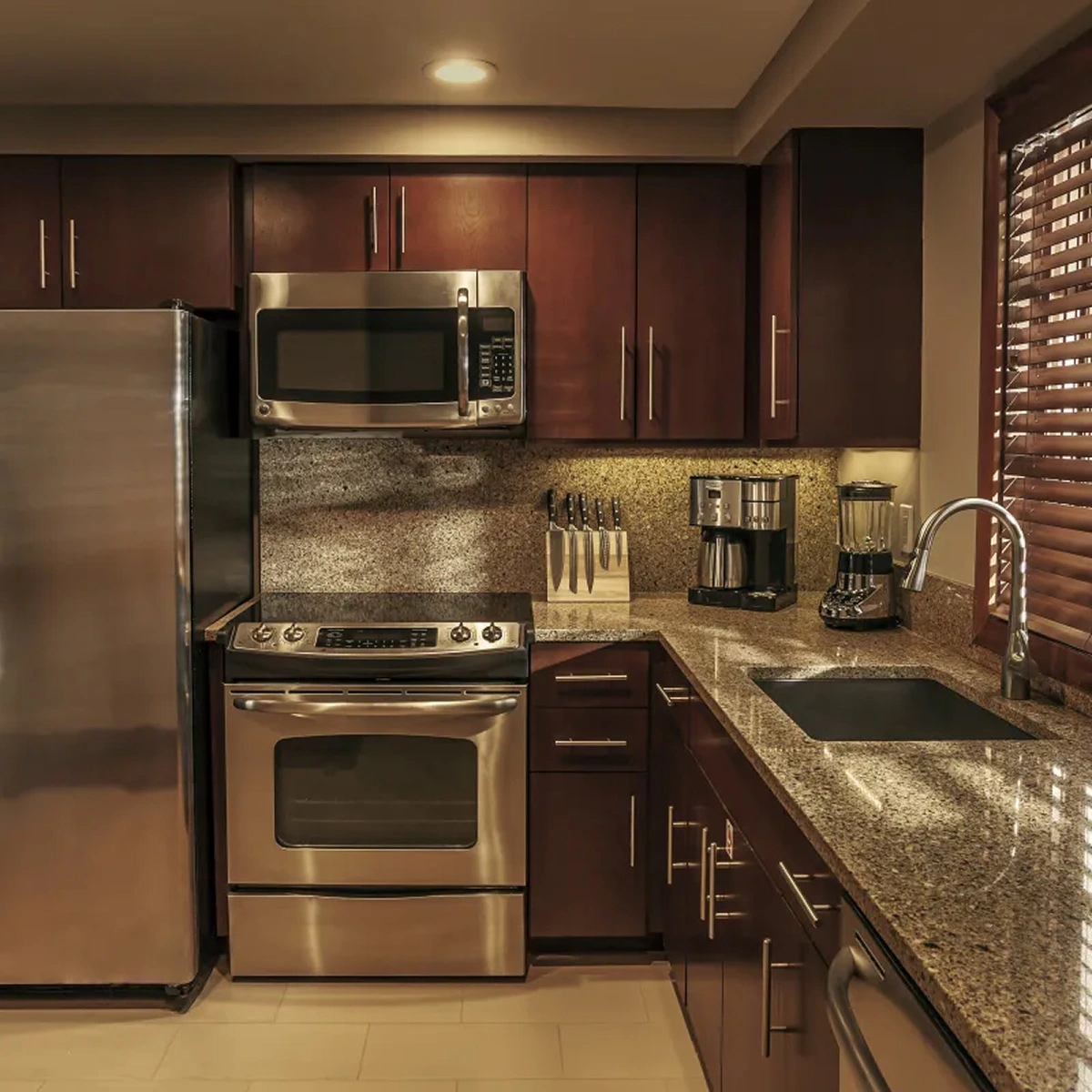 Modern kitchen with stainless steel fridge, oven, microwave, knife block, coffee maker, blender, and granite counters.