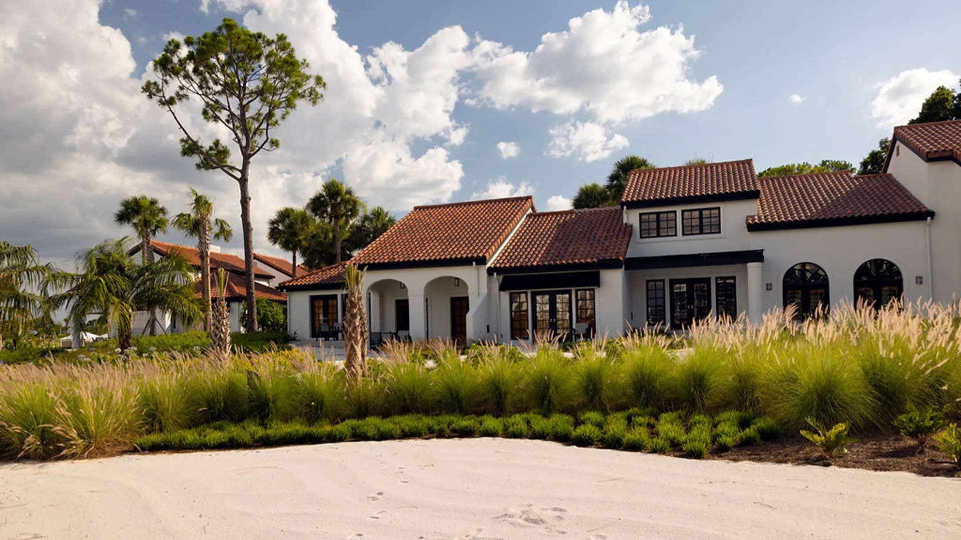 White villa with red tile roof behind tall grasses and palm trees under a partly cloudy sky.