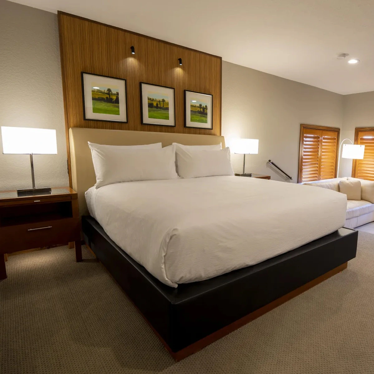 Modern hotel room with a large king bed, two bedside lamps, wall art, and a beige couch by shuttered windows.