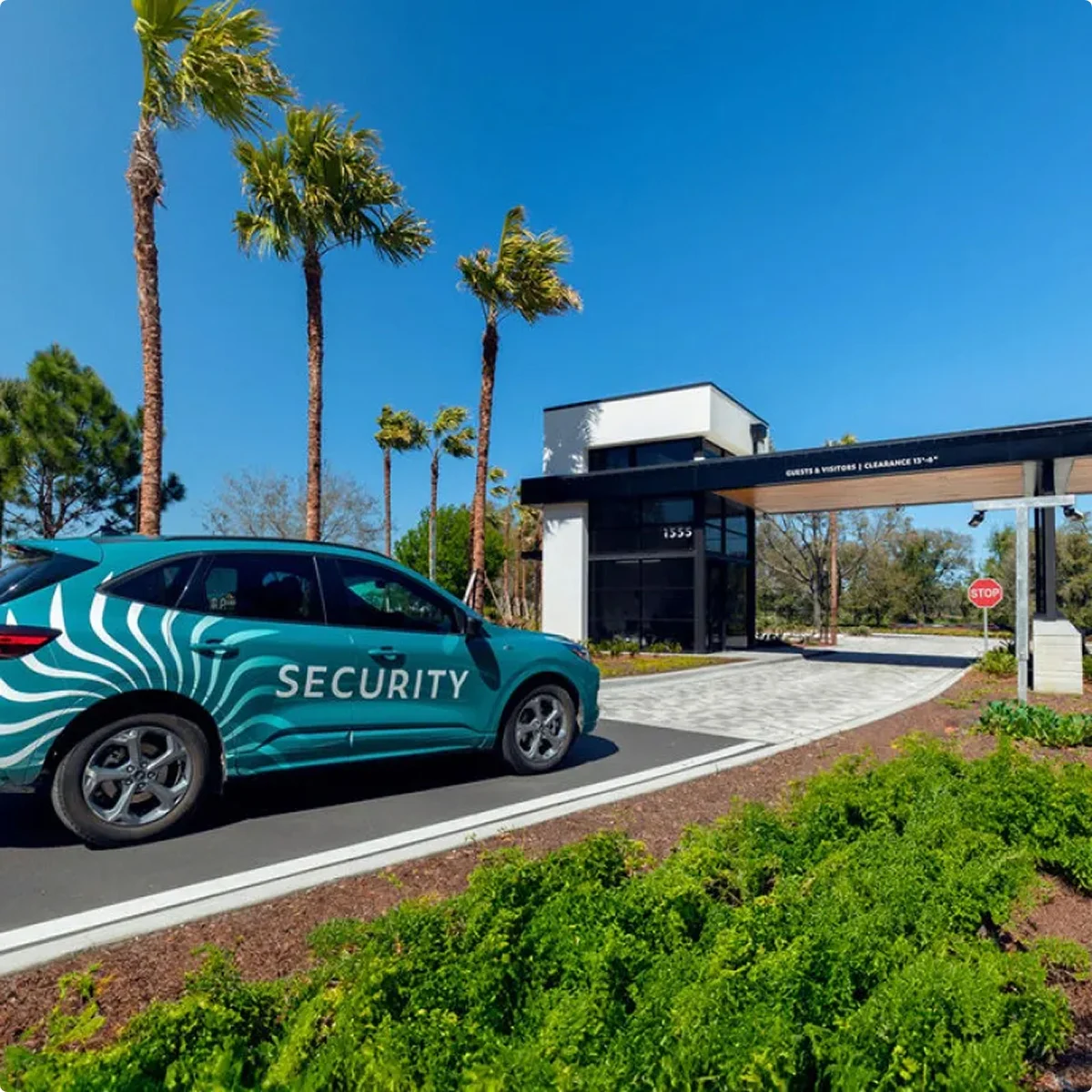 A teal security car parked near a gated entrance with palm trees and clear blue sky.