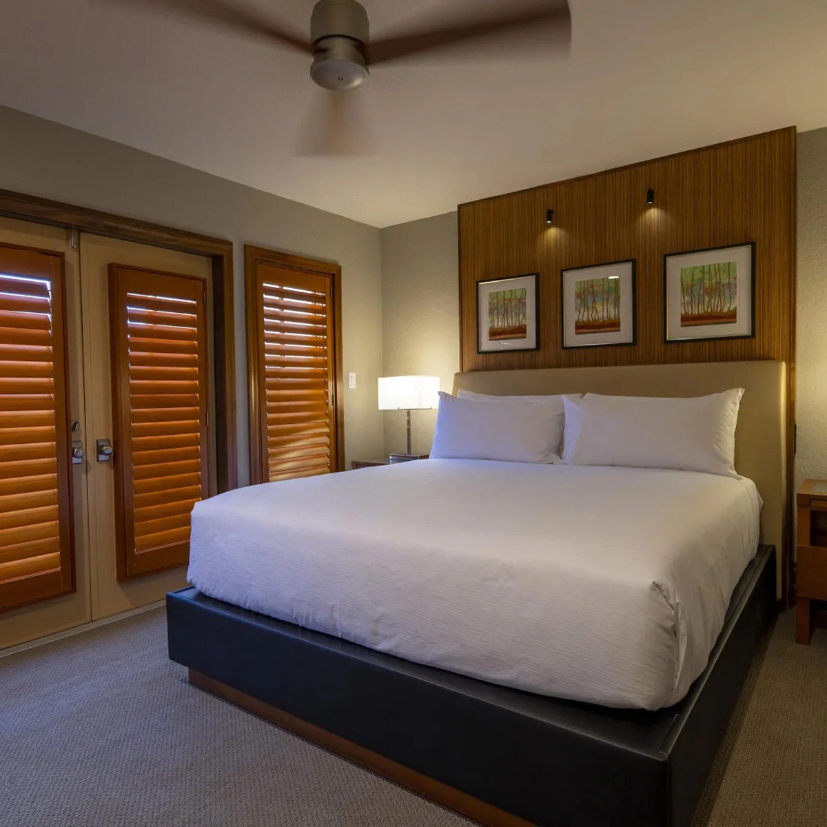 Modern bedroom with a large bed, white bedding, wood-paneled wall, framed art, and shuttered doors.