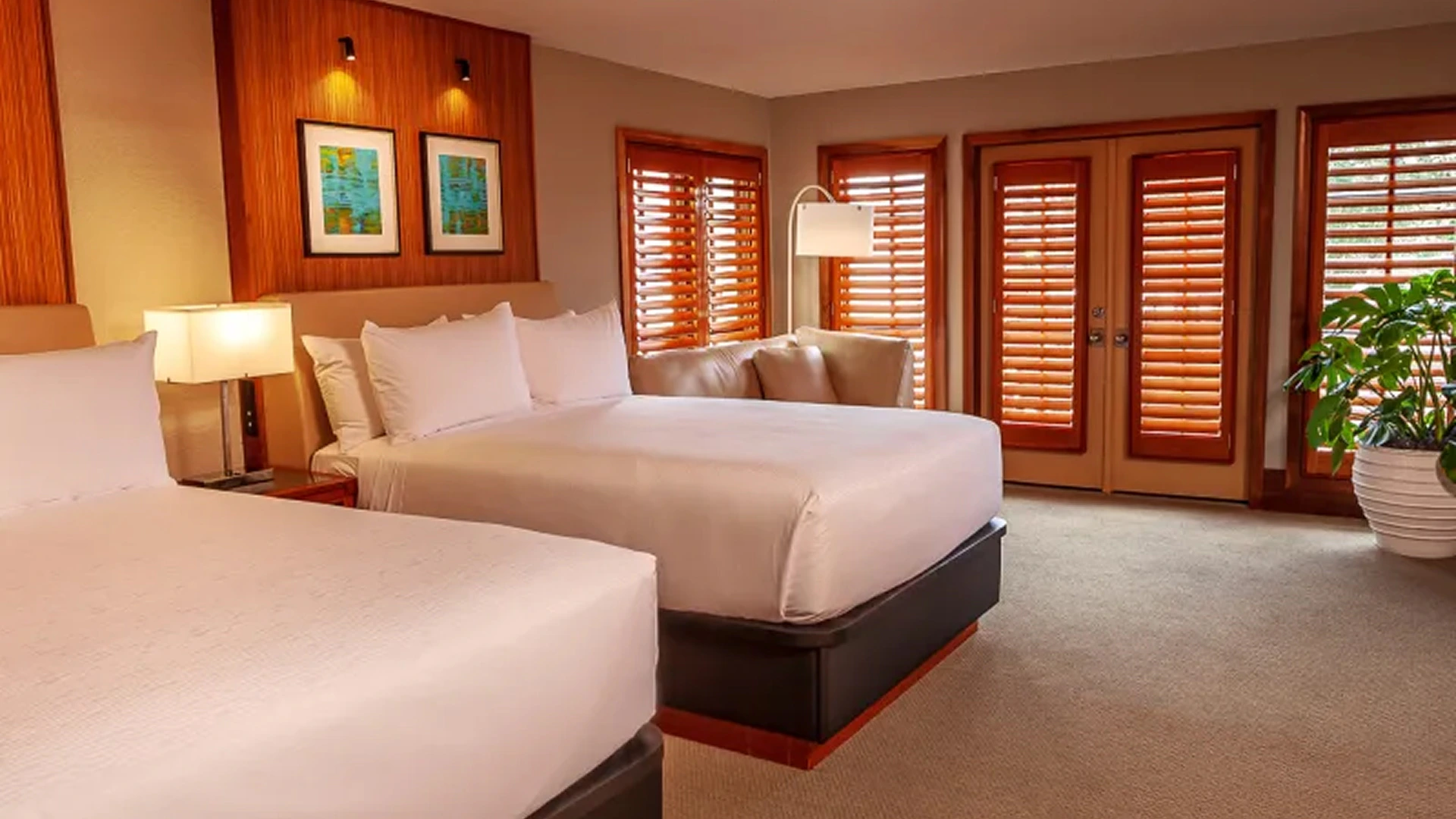 Hotel room with two queen beds, white linens, beige walls, wooden window shutters, and a potted plant.