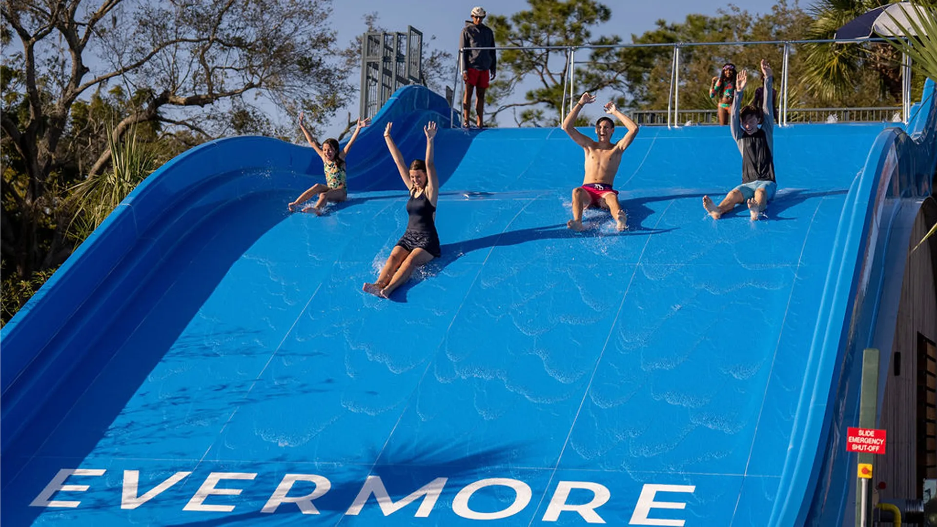 Four people sliding down a wide blue water slide with trees in the background.