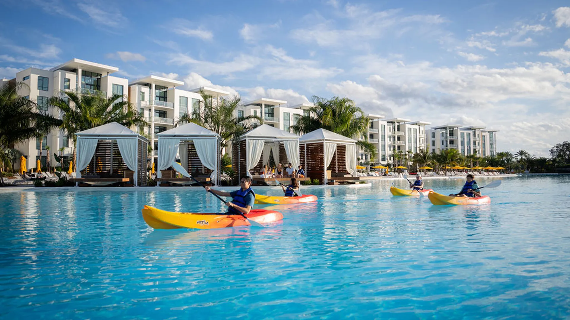 Four people kayaking in a large pool with cabanas and apartment buildings in the background.