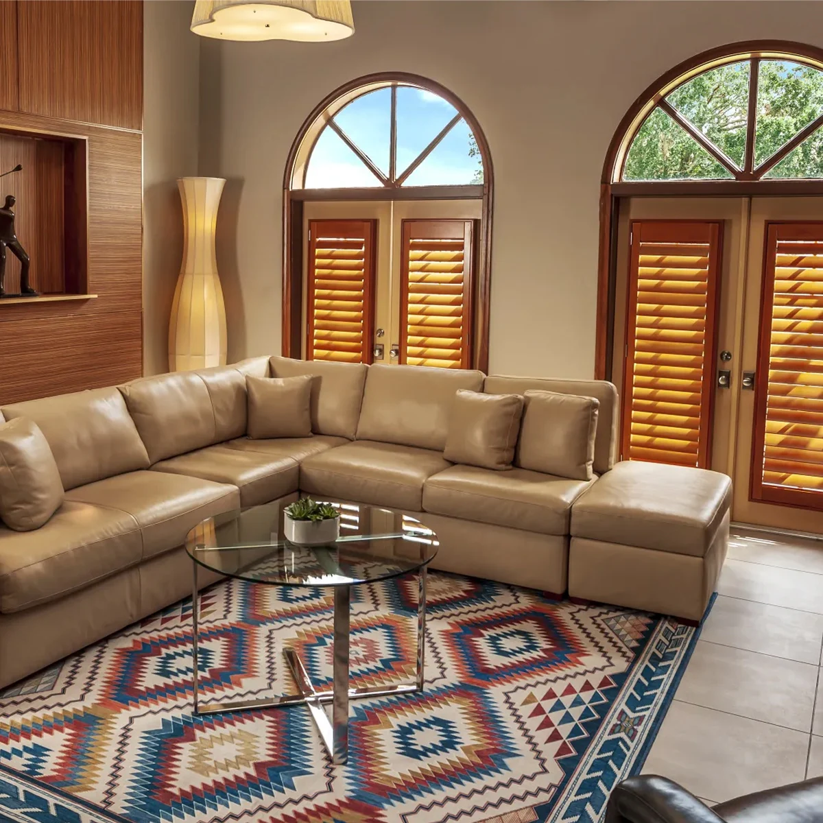 Living room with beige sectional sofa, glass coffee table, patterned rug, and arched windows with shutters.