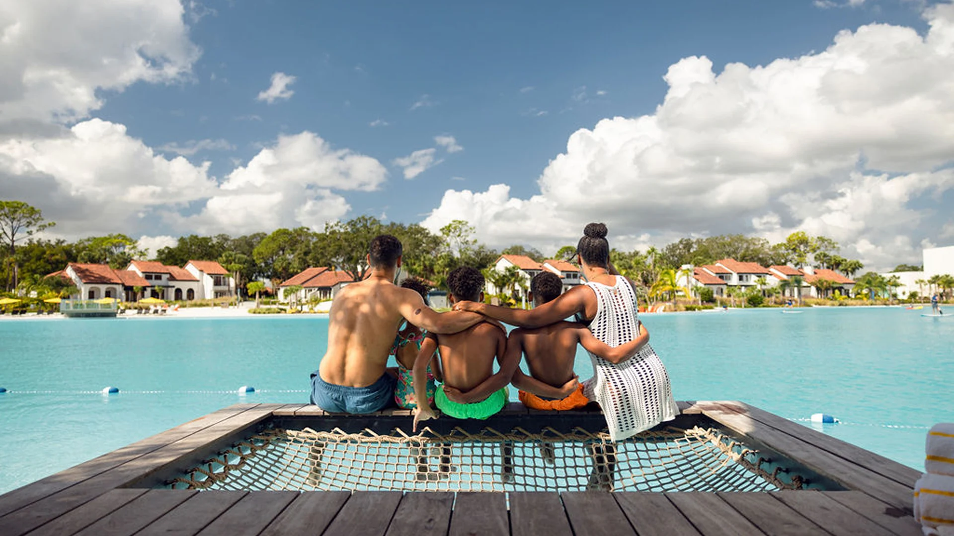 Five people sit side by side on a wooden dock by a blue lagoon, facing houses and trees.