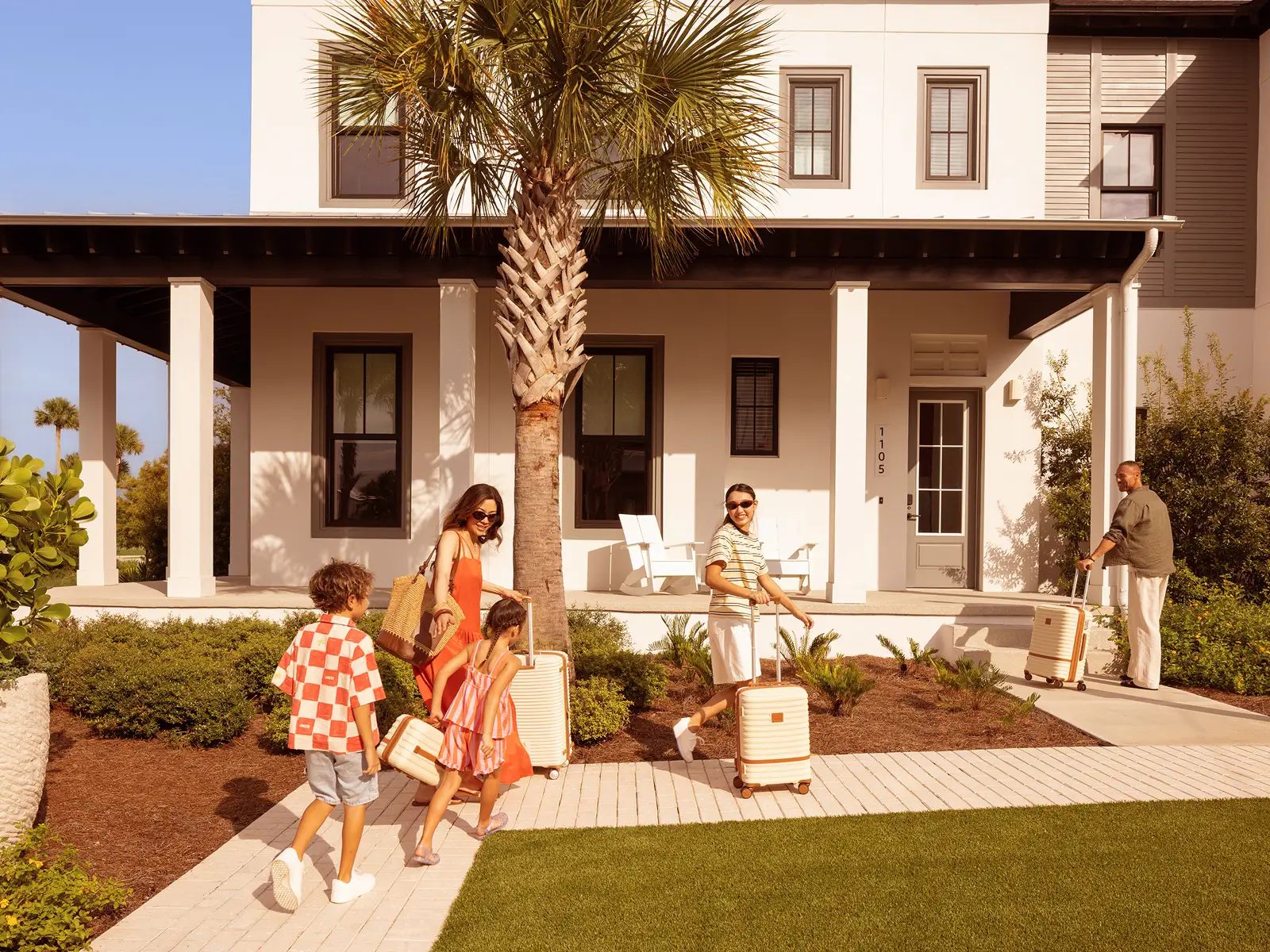 Family with luggage walking on sidewalk toward modern house with porch and palm tree.