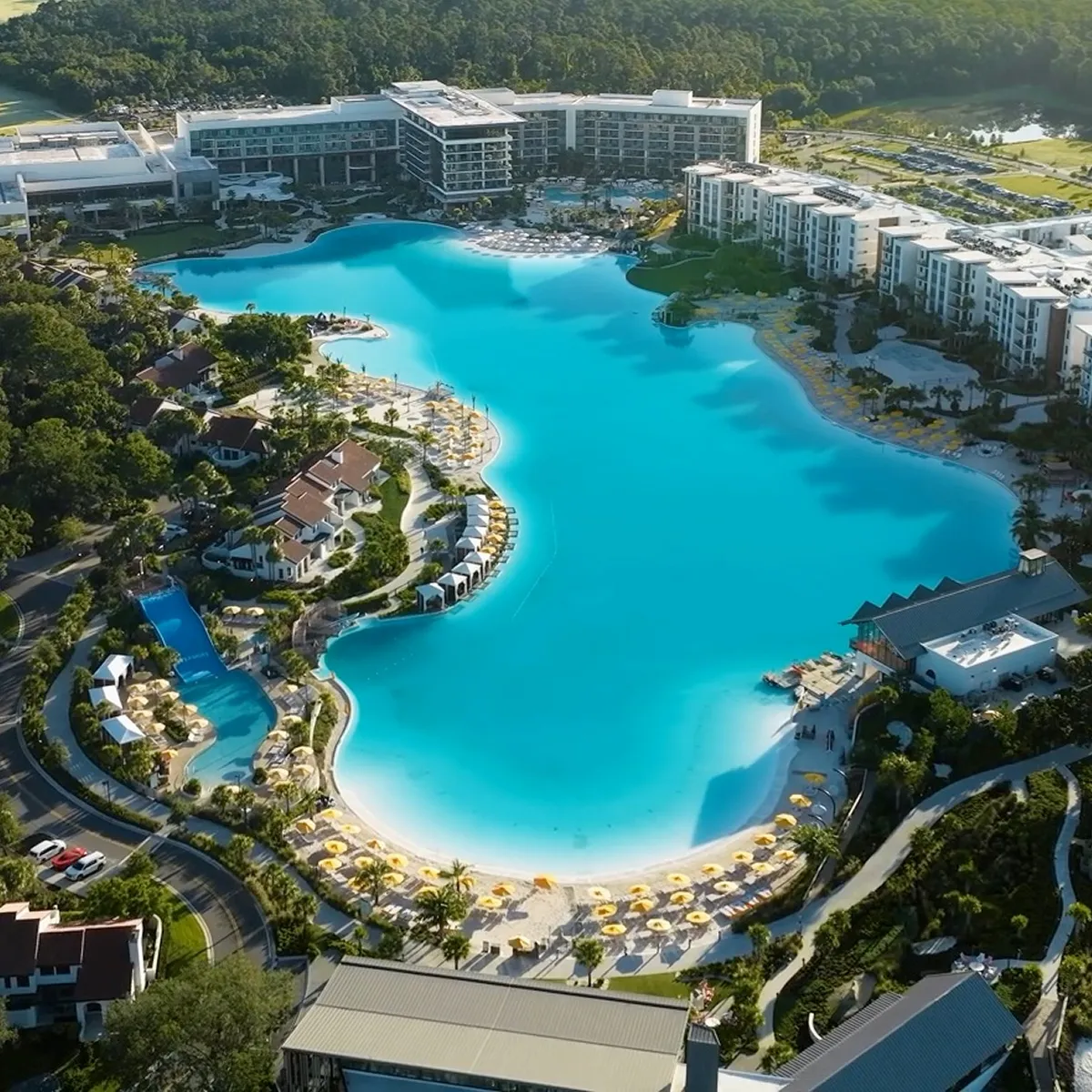 Aerial view of a large resort pool with yellow umbrellas, lounge chairs, and surrounding buildings in greenery.