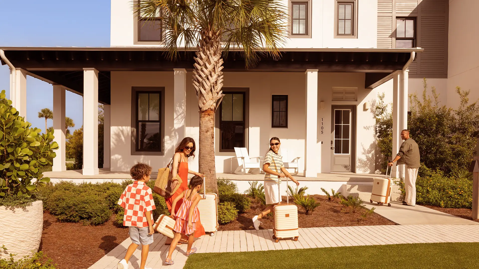 Family of five with suitcases walking toward a white two-story house with a front porch and palm tree.