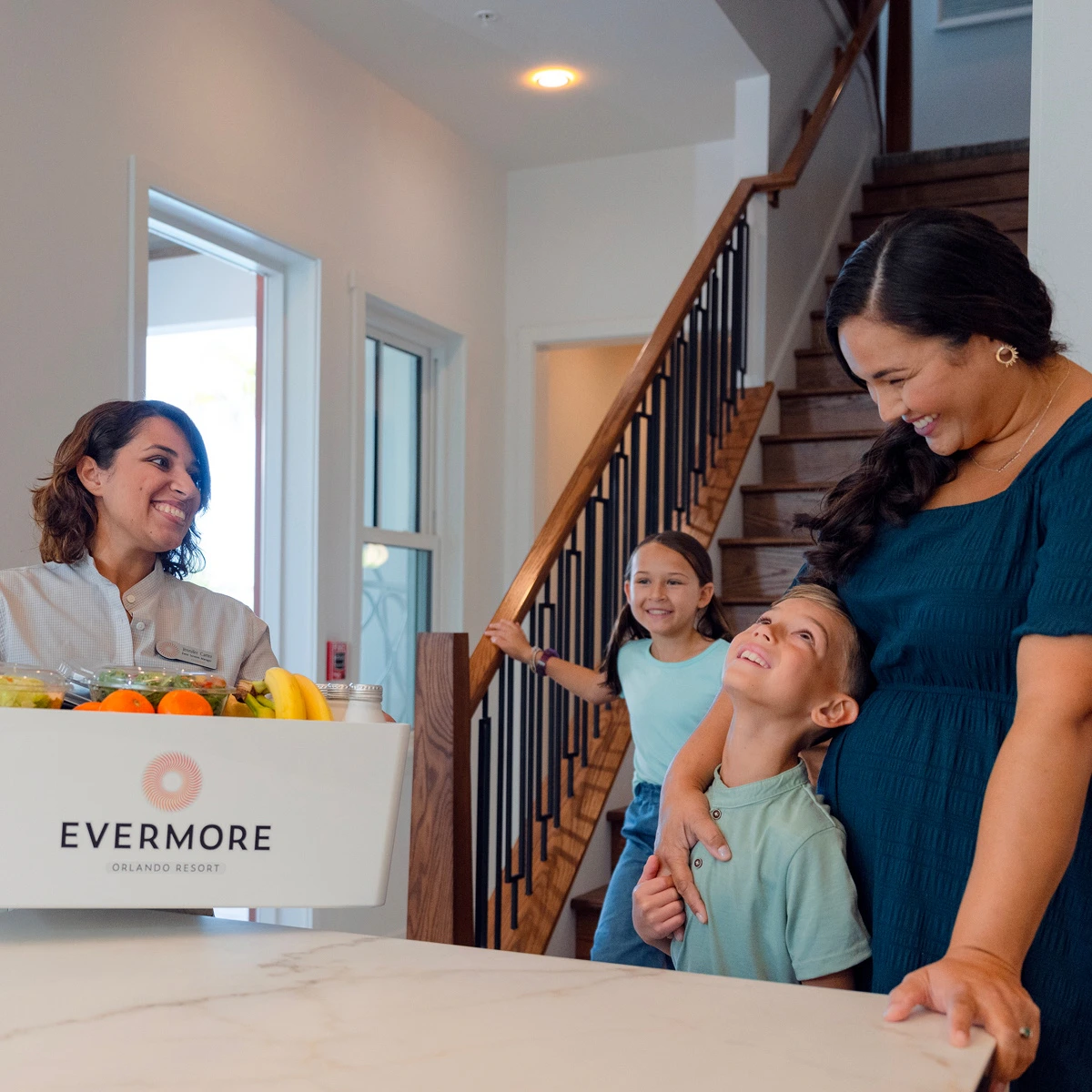 Woman delivering groceries to smiling family of three in a home with staircase in background.
