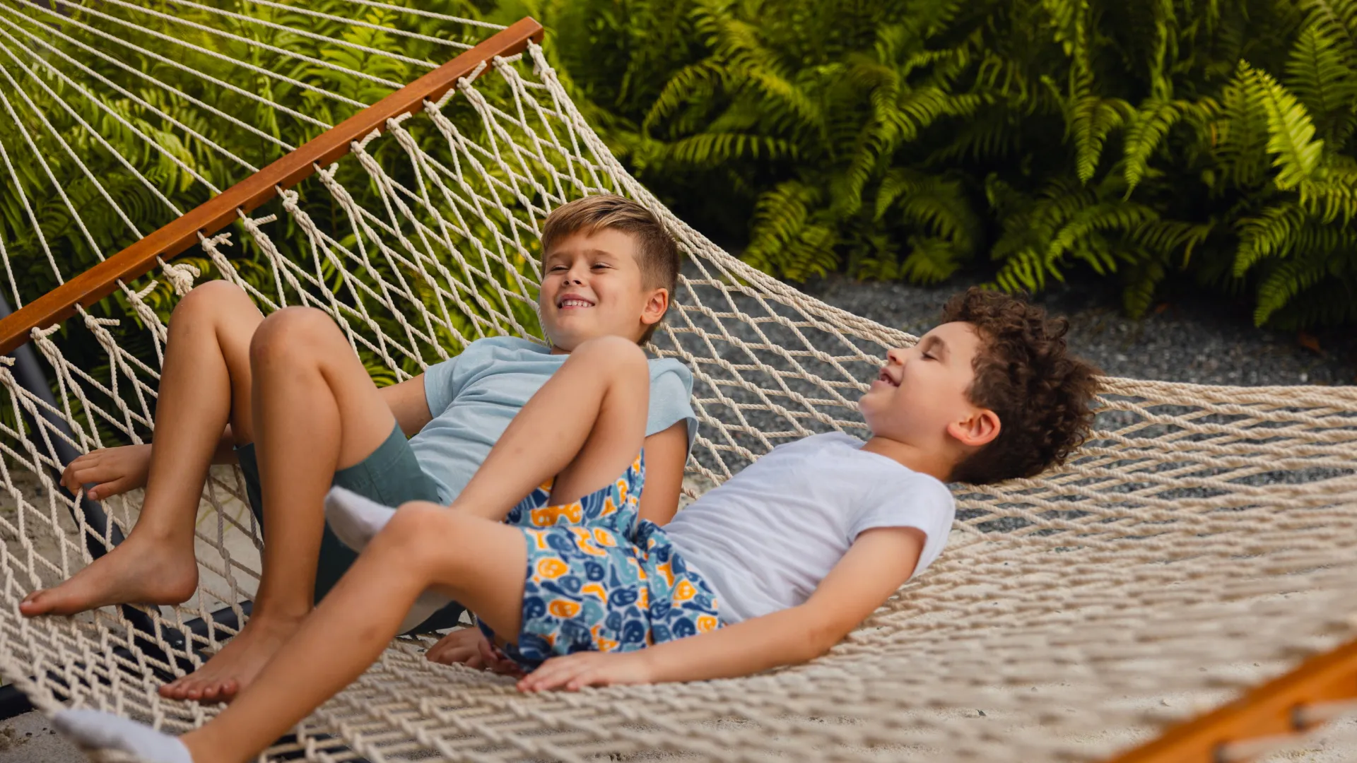Two boys lying and smiling together on a rope hammock outdoors near greenery.