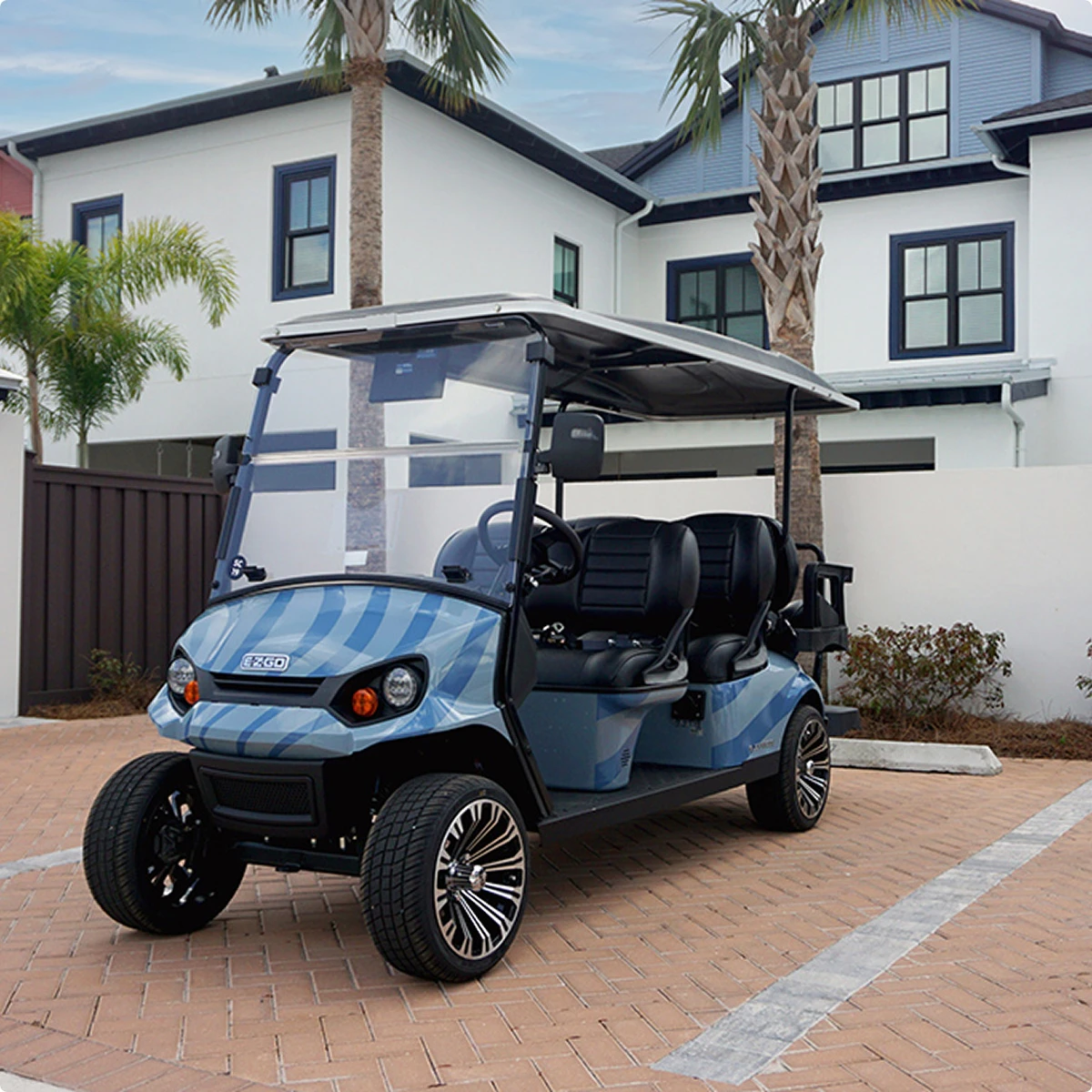 Blue and black golf cart parked on brick driveway in front of two-story houses and palm trees.