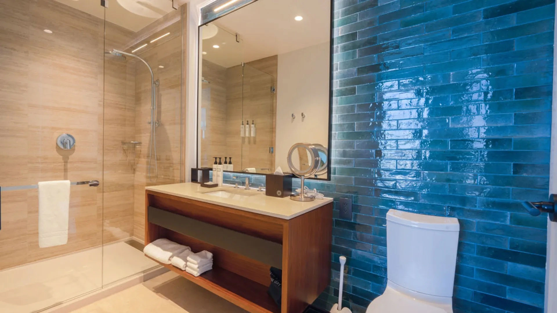 Modern bathroom with blue tiled wall, wooden vanity, white toilet, and glass-enclosed shower.