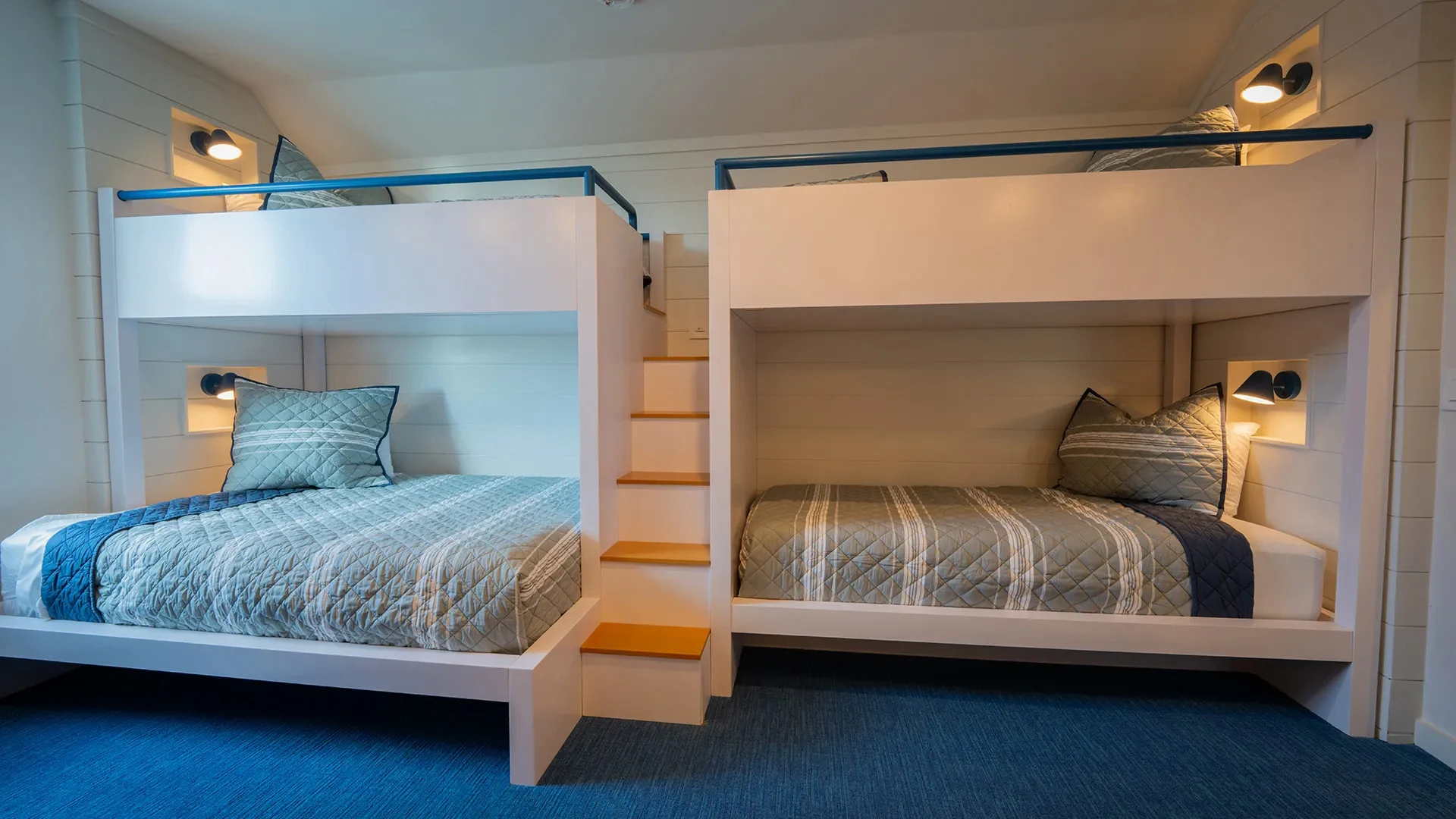 Modern white bunk beds with blue-gray quilts and pillows, with wooden steps in the center.