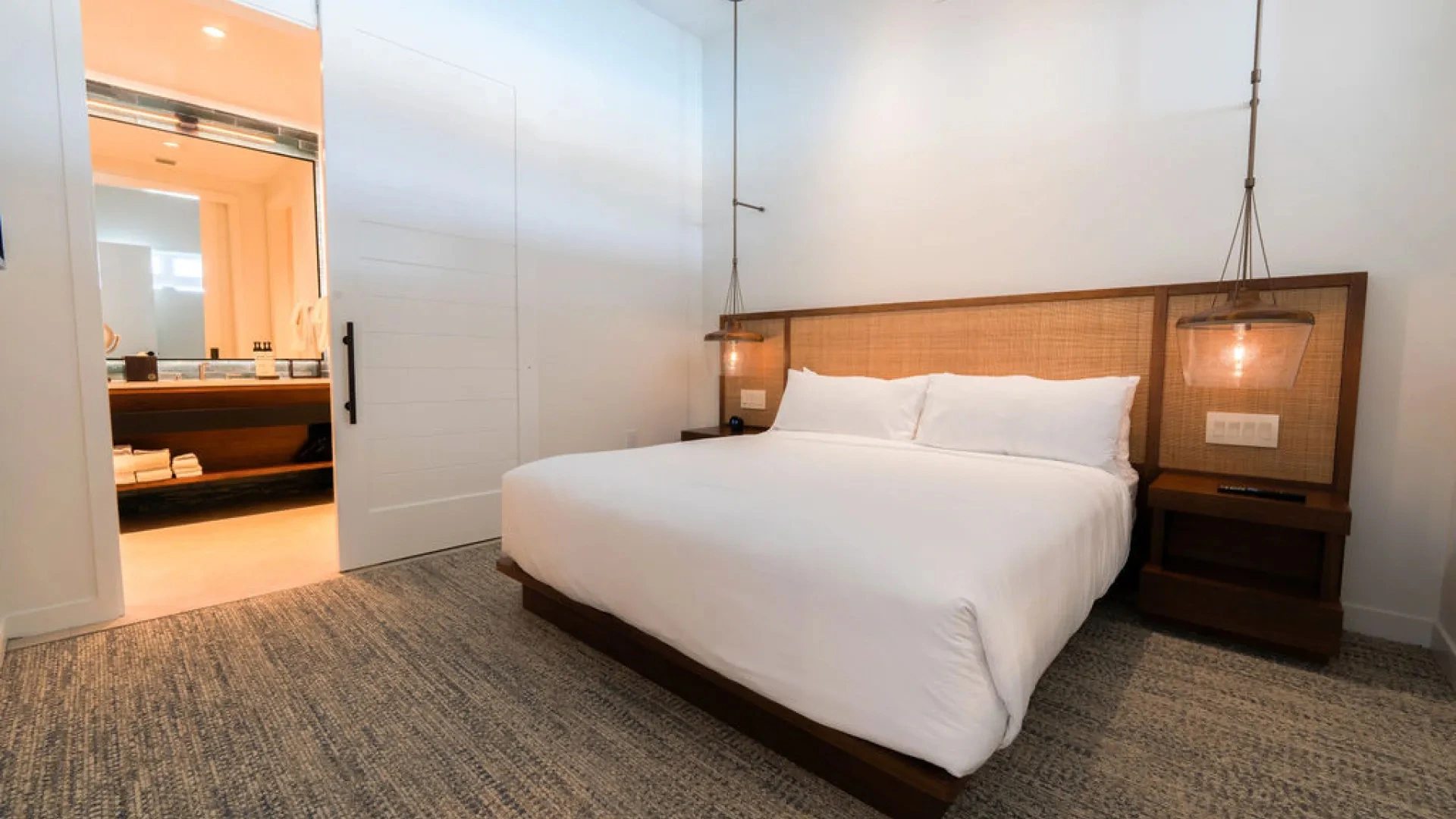 Modern bedroom with a white bed, wooden headboard, hanging lights, and open door to bathroom.