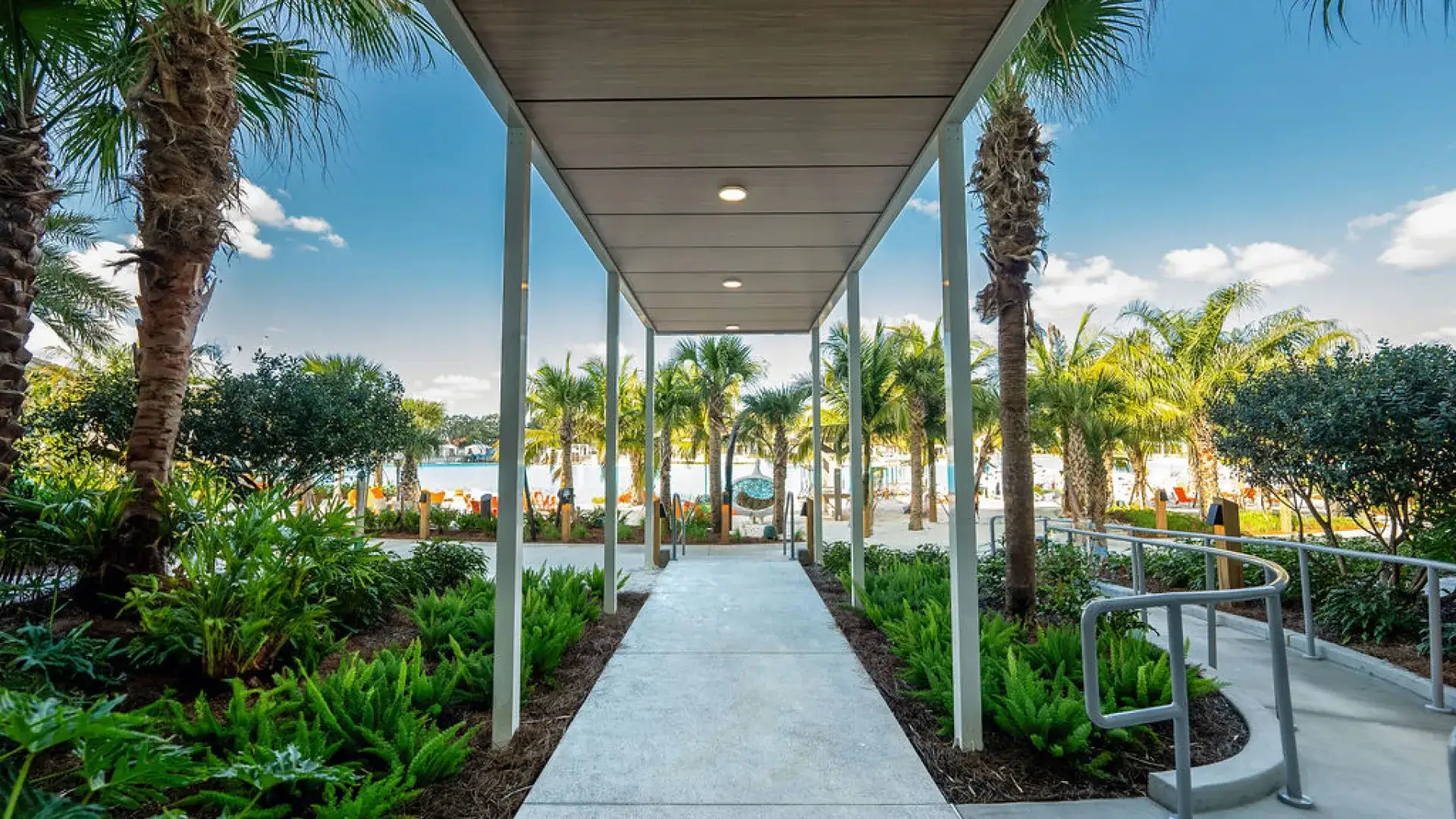Covered walkway flanked by palm trees and greenery leads to a sunny beach area with lounge chairs.