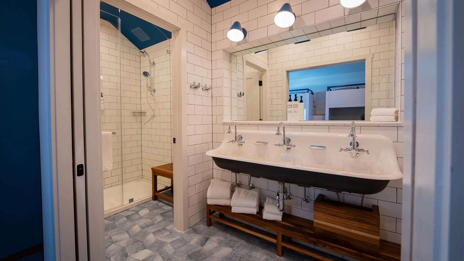 Bathroom with triple sink, large mirror, white tile walls, and a glass-enclosed shower with wooden bench.