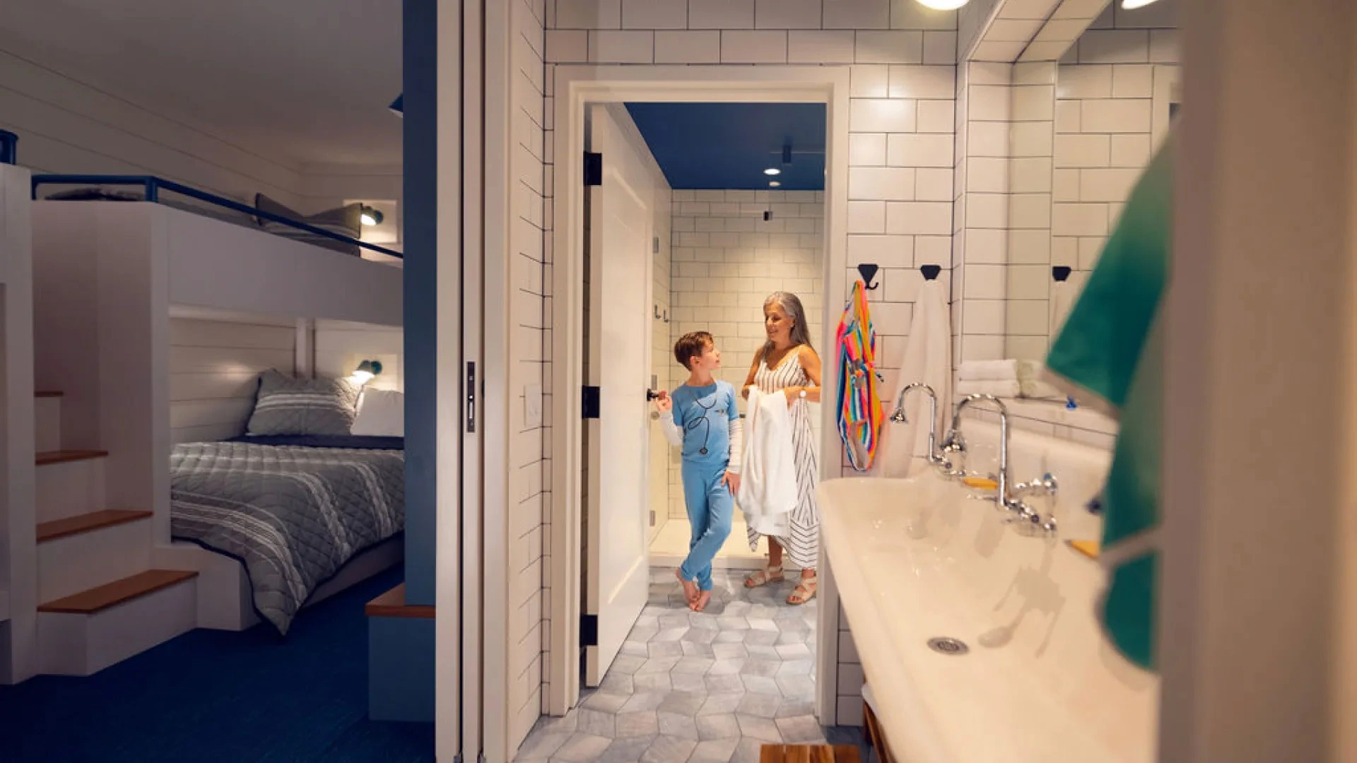 Child in blue pajamas and woman with towel standing in bathroom doorway beside sink and bedroom.