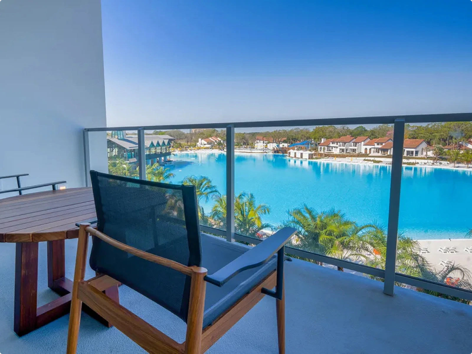 Balcony with a wooden table and chair overlooking a large blue pool and houses with red roofs.