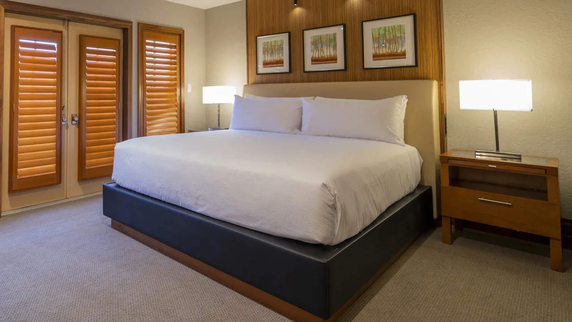 Modern bedroom with king-size bed, white bedding, wood-paneled wall, three framed pictures, and bedside lamps.