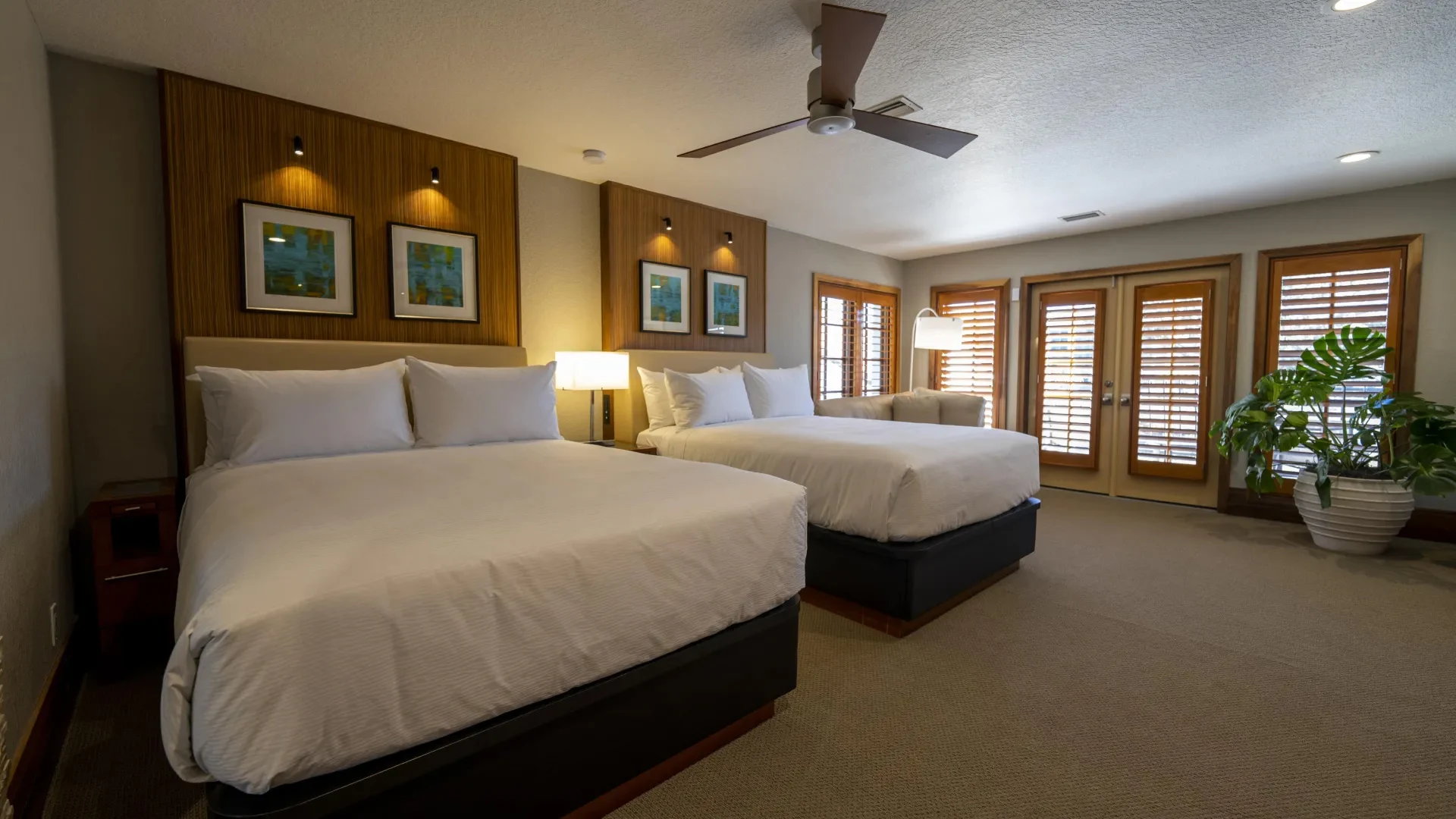 Modern hotel room with two large beds, bedside lamp, armchair, and windowed doors with shutters.