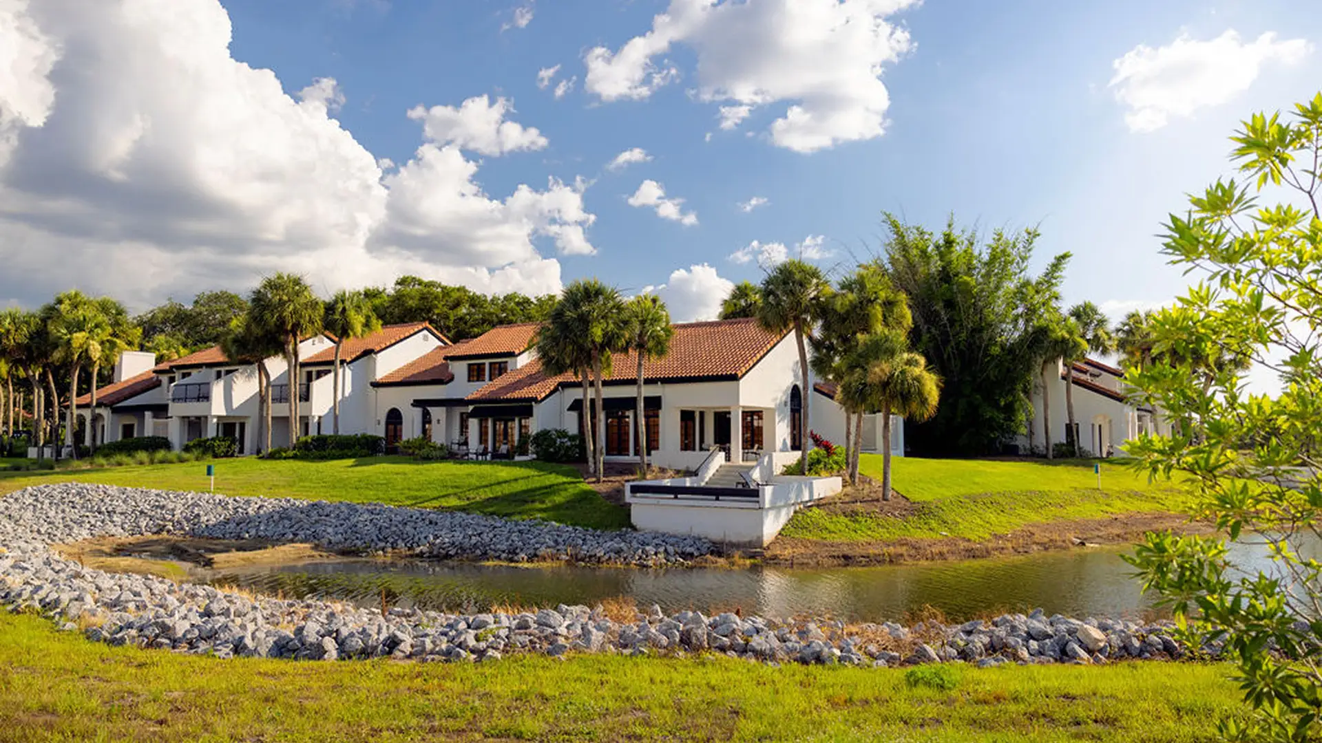White villas with red-tile roofs by a small water body, surrounded by palm trees and green grass.