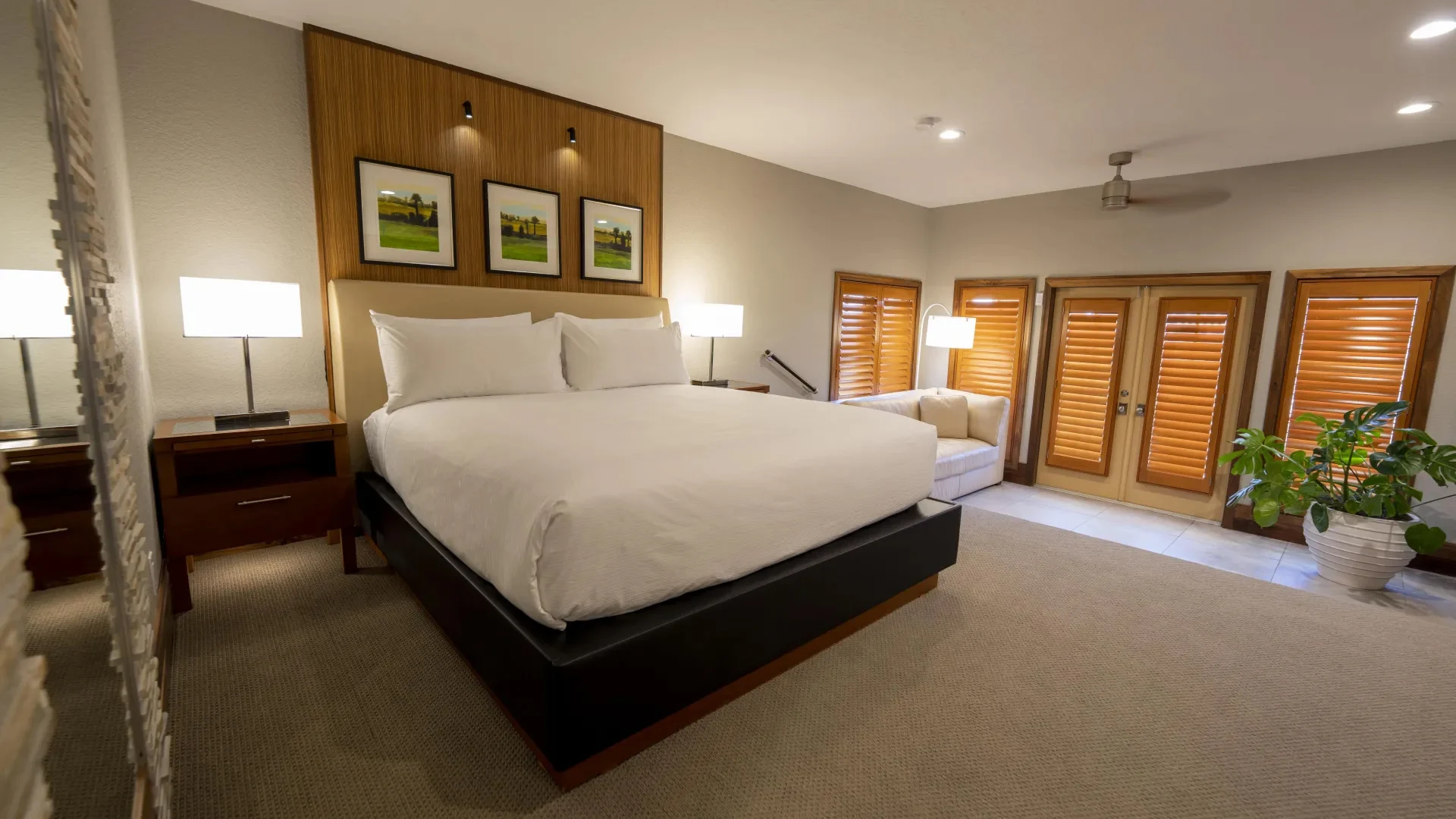 Modern bedroom with king-size bed, bedside lamps, sofa near wooden shuttered windows, and potted plant.