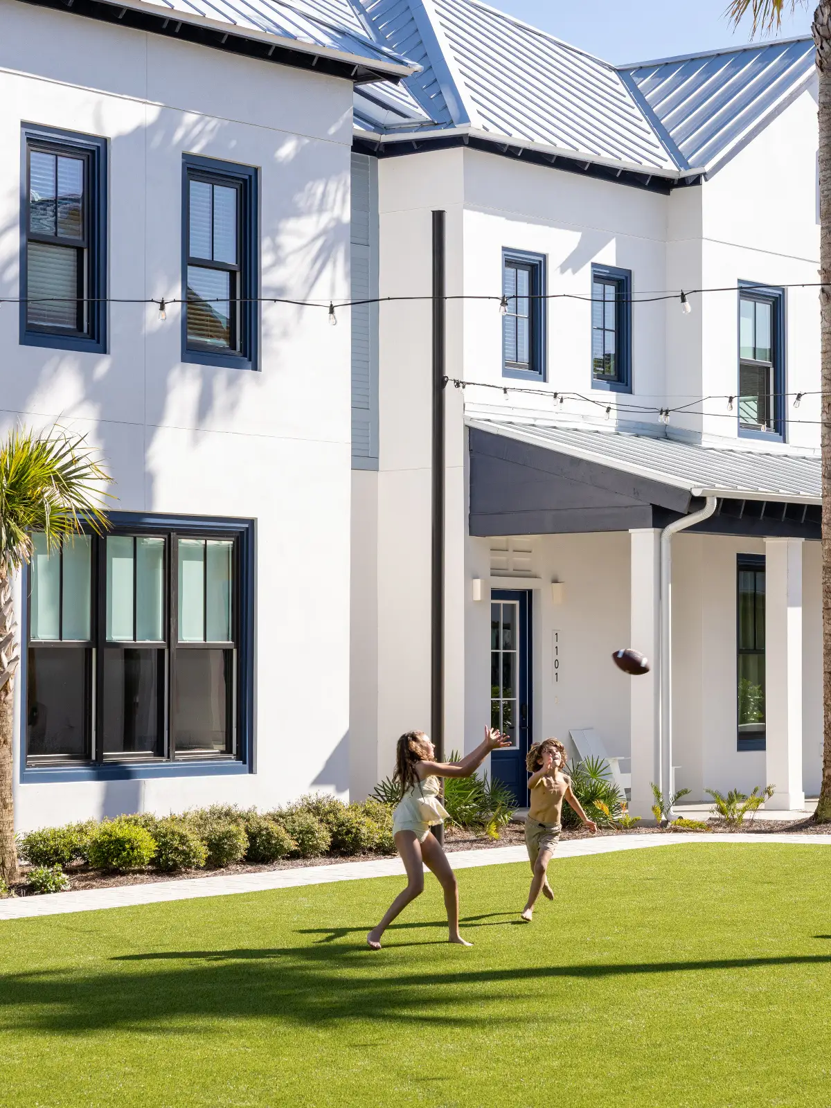 Two children playing catch with a football on a green lawn in front of a white house.