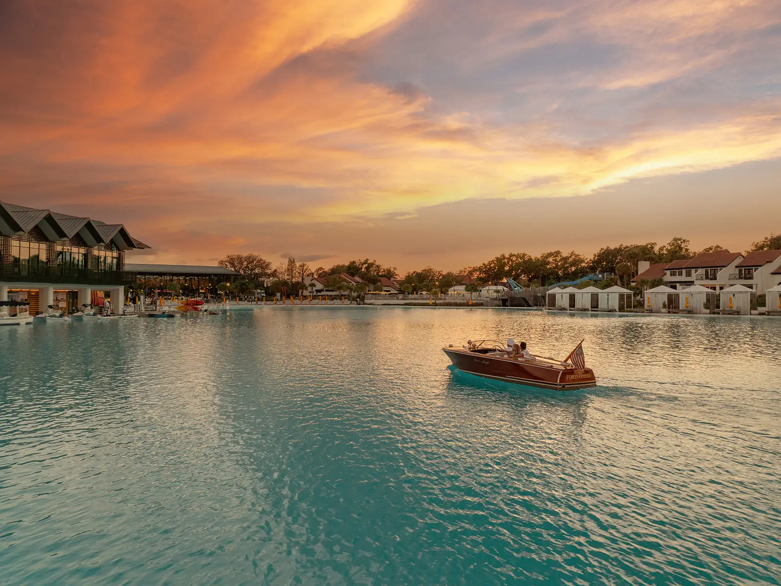 Small wooden boat with people cruises on a large lagoon near resorts at sunset with colorful sky.