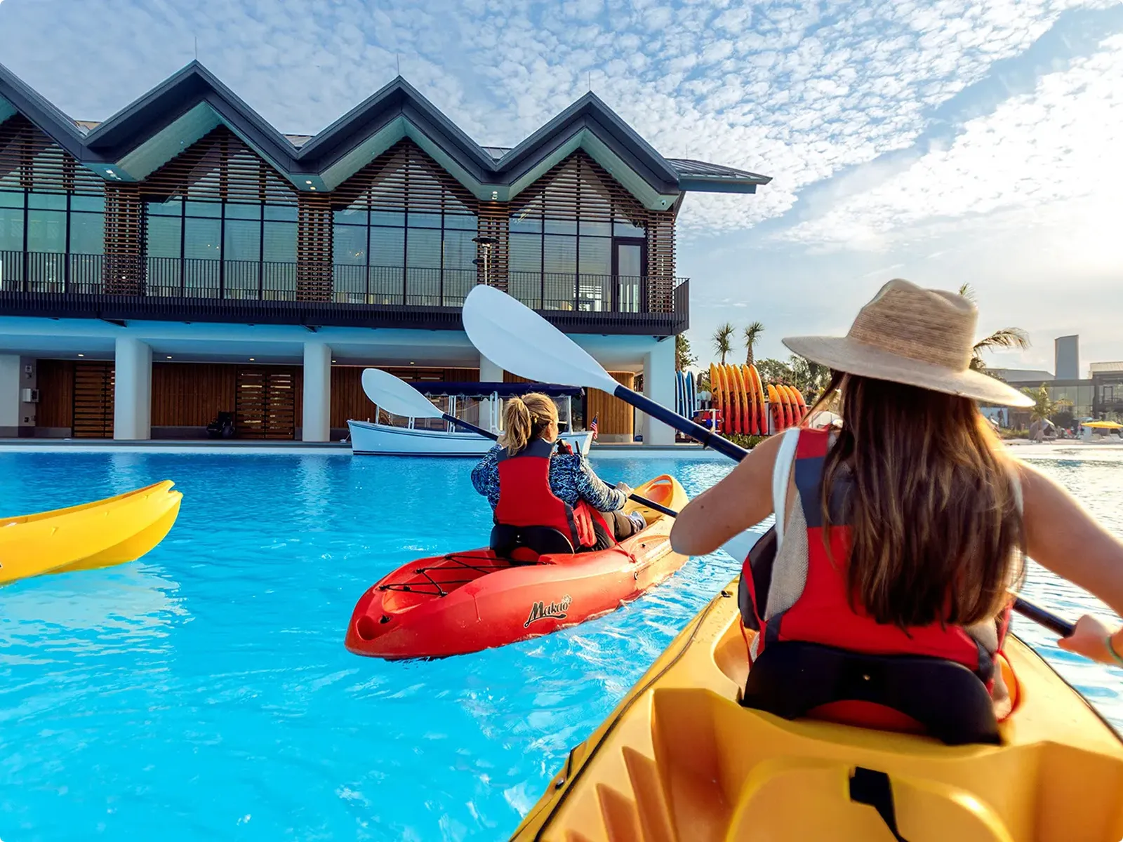Two people kayaking in a pool near a modern building under a blue sky with scattered clouds.