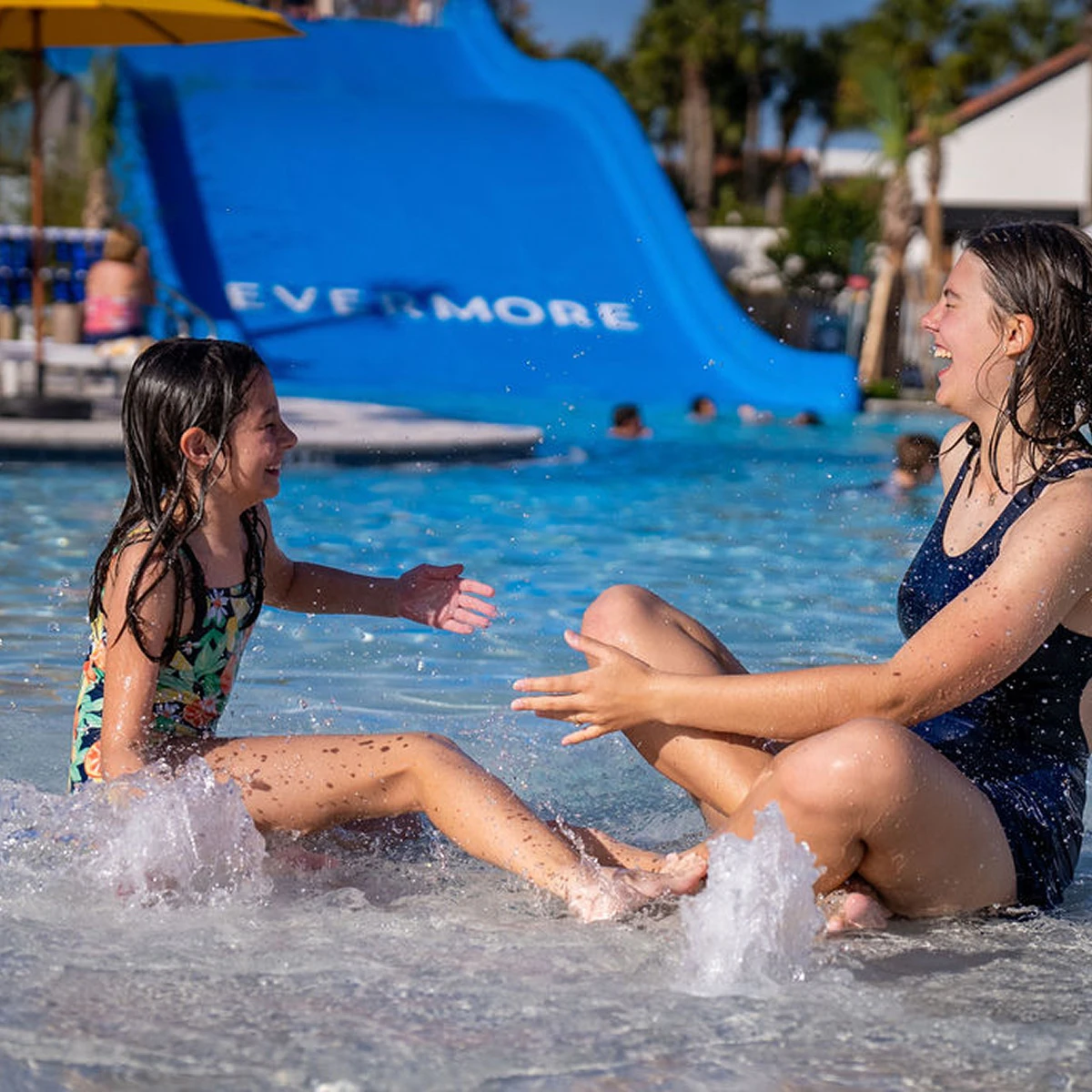 Two girls laughing and splashing water while sitting in a shallow pool near a blue water slide.