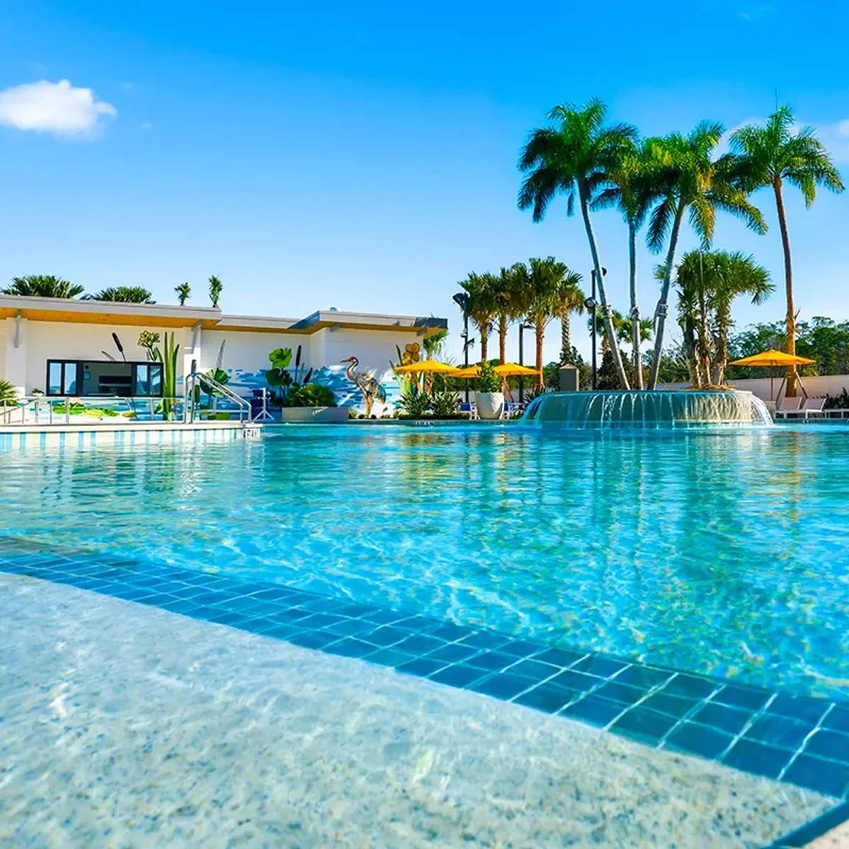 Clear outdoor pool with fountain, palm trees, yellow umbrellas, and building with nature mural in background.