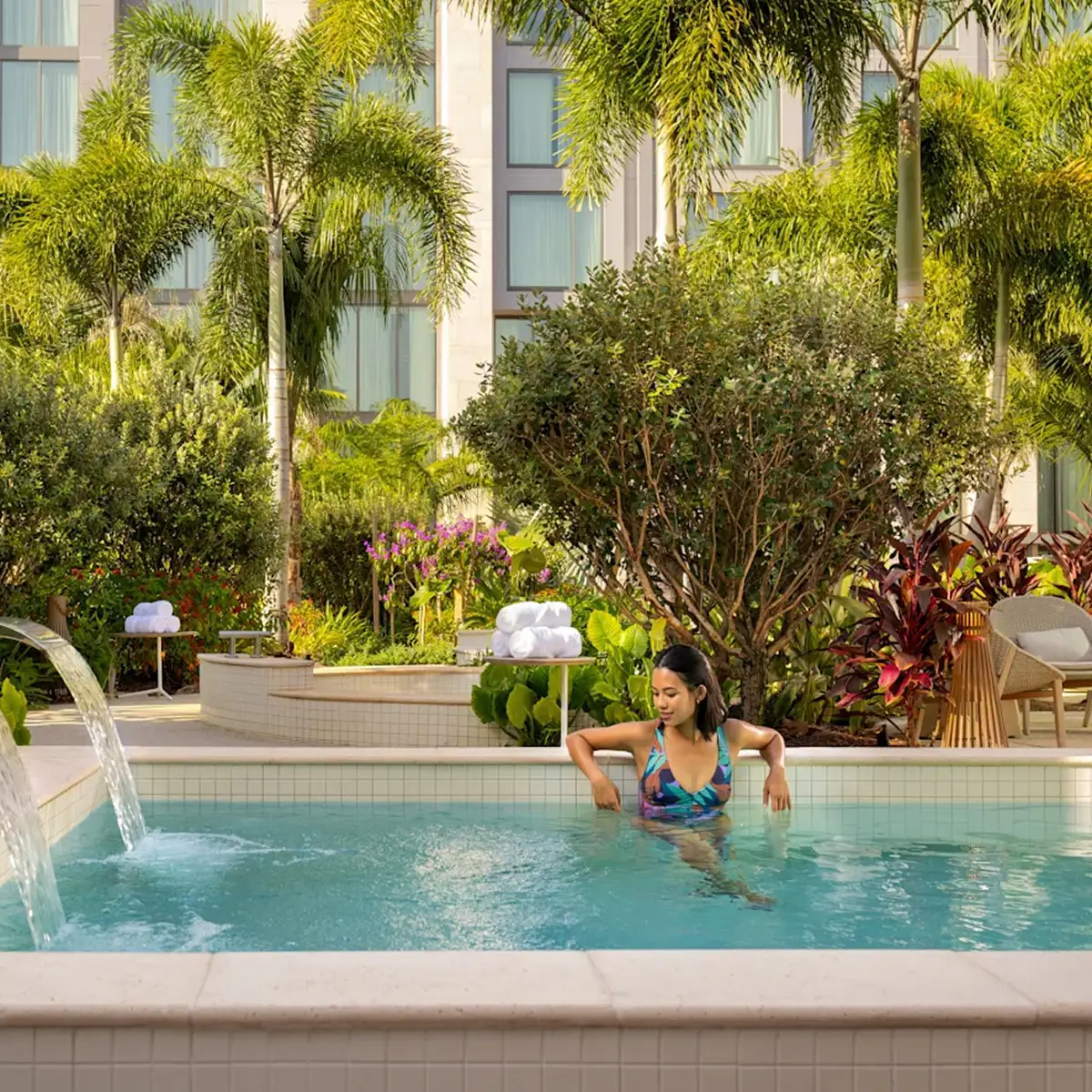 Woman relaxing in a tiled spa pool surrounded by tropical plants and palm trees.