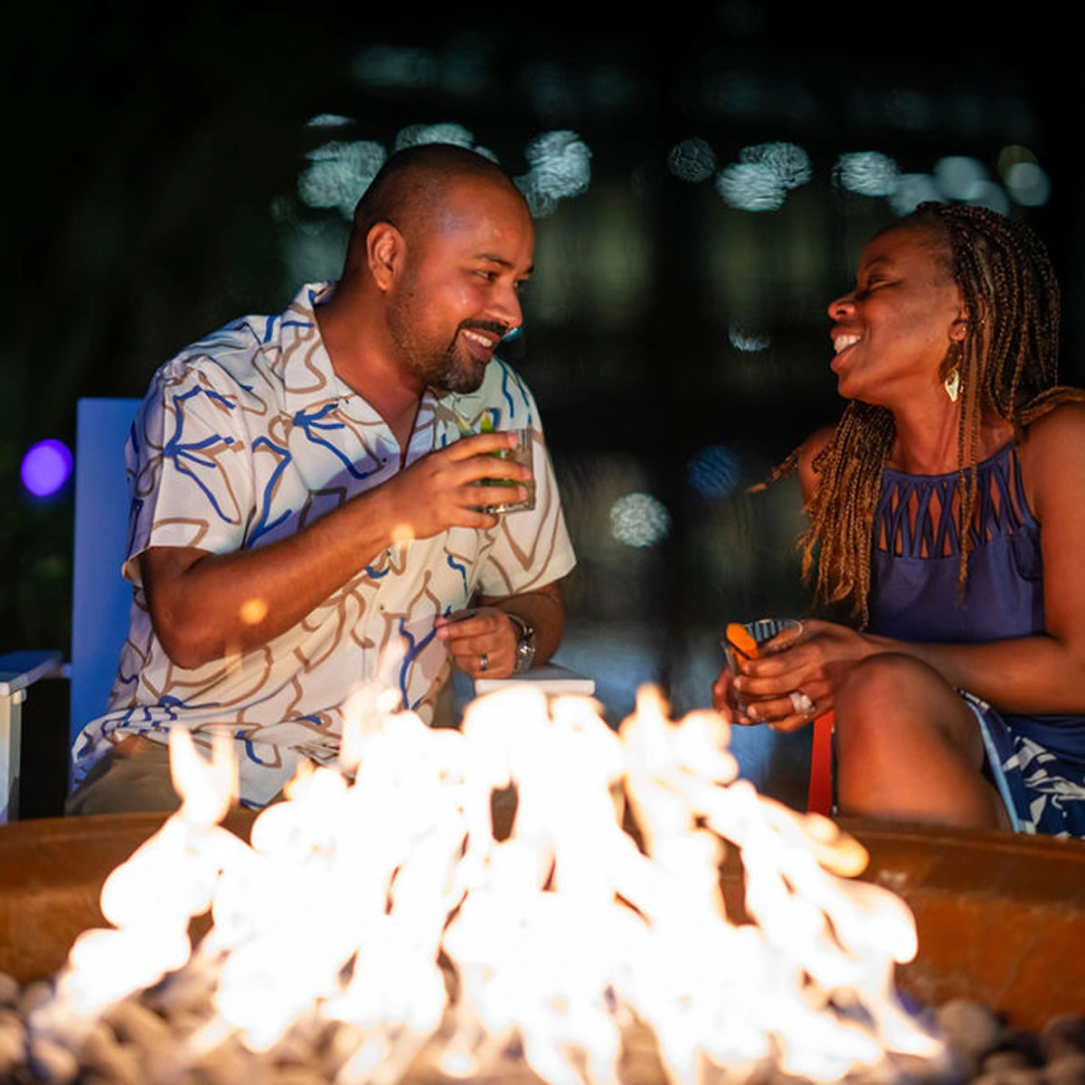 Man and woman smiling and chatting by a fire pit at night, holding drinks and sitting on chairs.